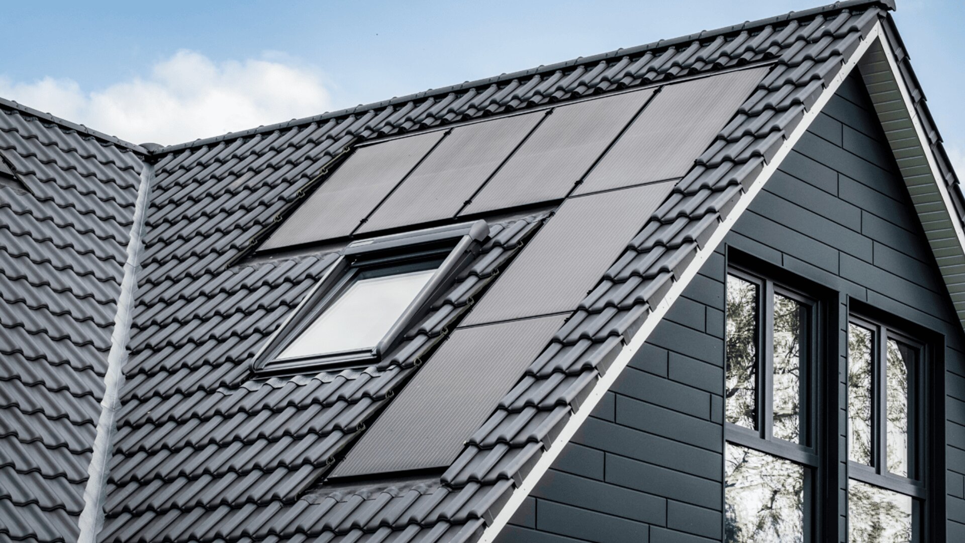 Auf einem Schrägdach mit dunkelgrauen Ziegeln befinden sich ein modernes Oberlichtfenster und eine Reihe schwarzer, in das Dach integrierter Solarzellen. Der Himmel im Hintergrund ist teilweise bewölkt.