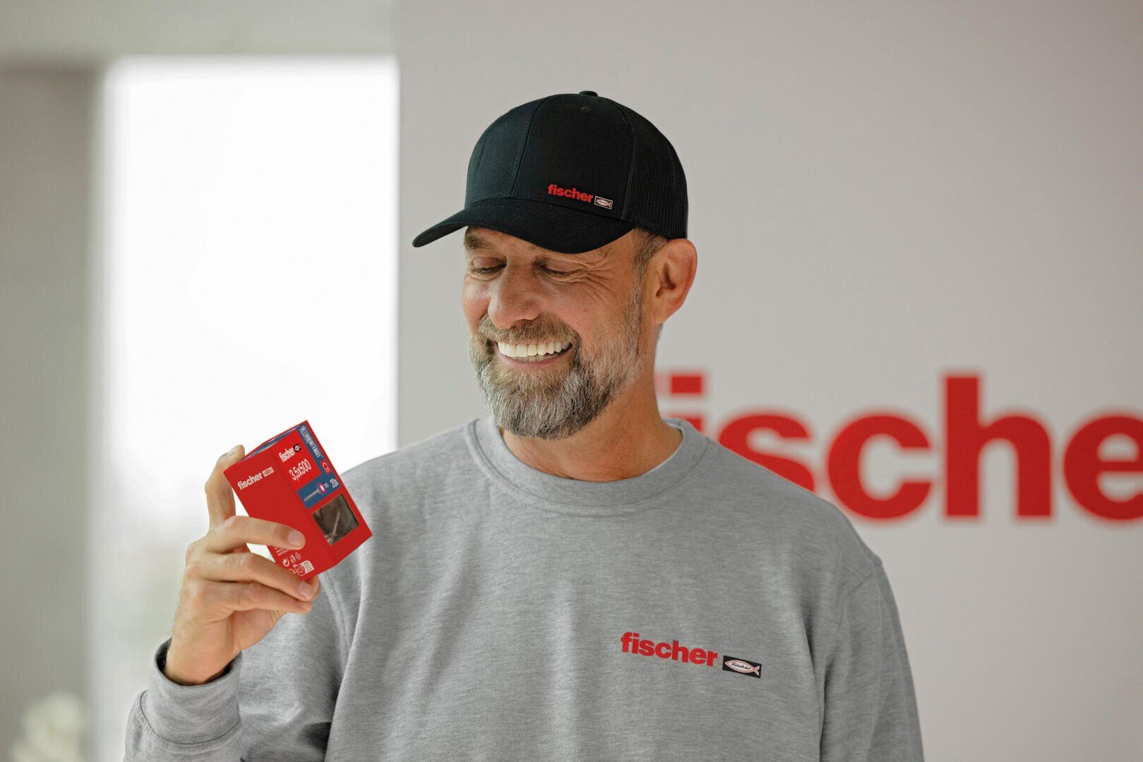 Ein lächelnder Mann mit Fischer-Mütze und Sweatshirt hält eine kleine rote Schachtel. Im Hintergrund ist eine unscharfe Wand zu sehen, auf der in roten Buchstaben teilweise das Wort fischer zu lesen ist.