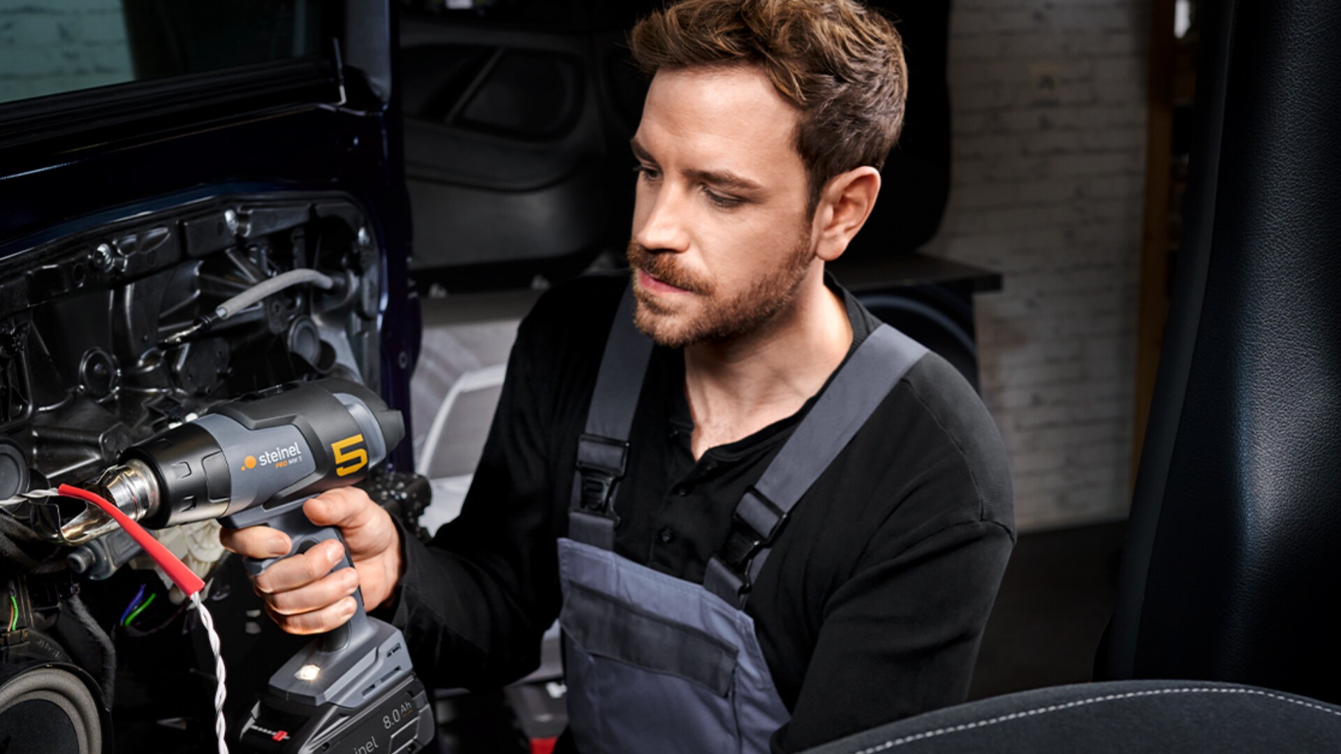 Ein Mann im Overall arbeitet mit einer Bohrmaschine an der Verkabelung im Inneren einer Autotür und scheint sich auf die Reparaturaufgabe zu konzentrieren.