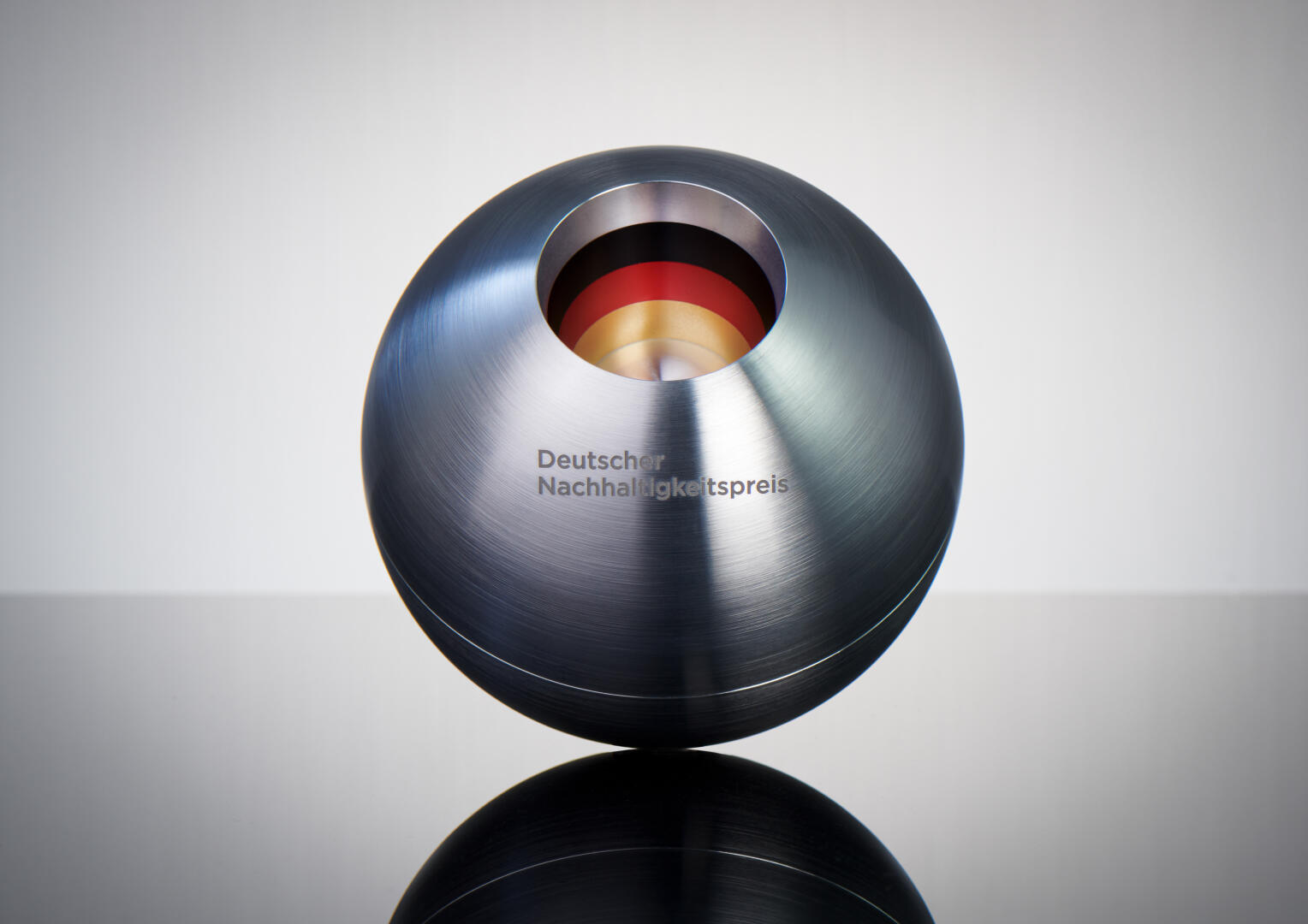 Eine metallische, kugelförmige Trophäe mit offener Oberseite, die im Inneren konzentrische farbige Ringe freigibt, in die die Worte „Deutscher Nachhaltigkeitspreis“ eingraviert sind und die auf einer reflektierenden Oberfläche präsentiert werden.