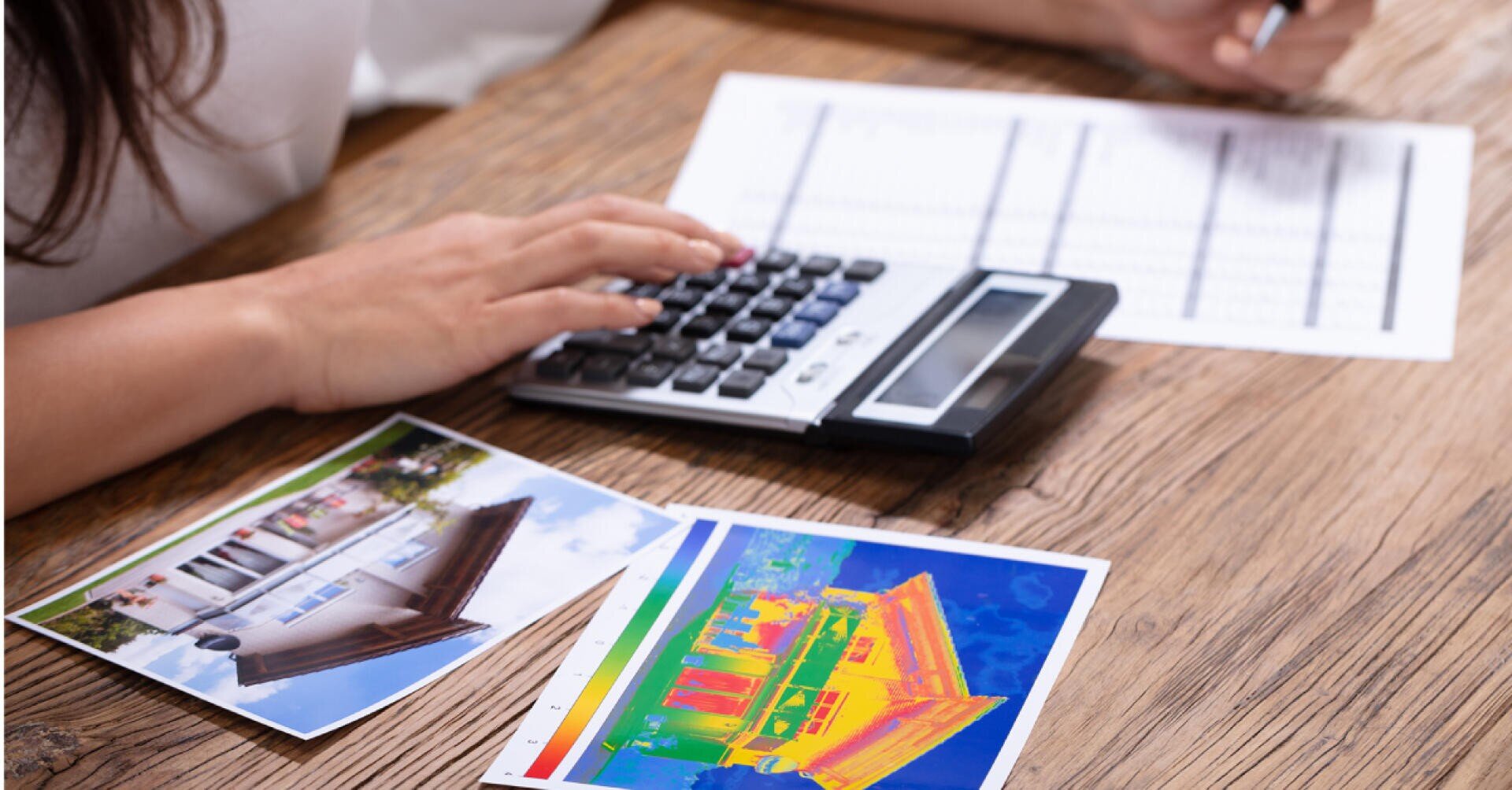Eine Person benutzt einen Taschenrechner neben einem Dokument und zwei Fotos, eines von einem Haus und eines mit einer Wärmebildaufnahme eines Gebäudes, auf einem Holztisch.