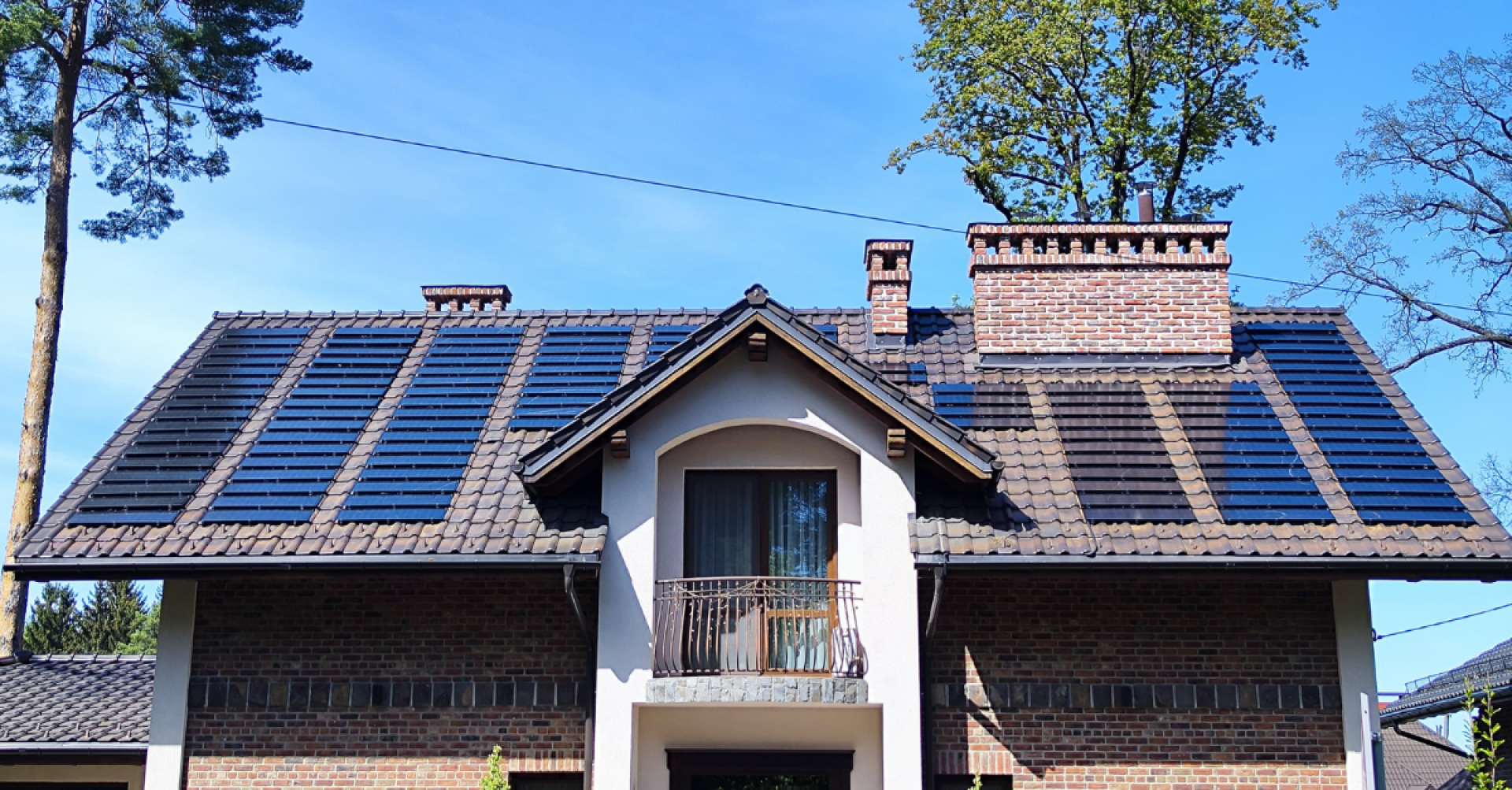 Ein Haus mit einem Ziegeldach mit integrierten Sonnenkollektoren und zwei großen Backsteinschornsteinen vor blauem Himmel und Bäumen im Hintergrund.