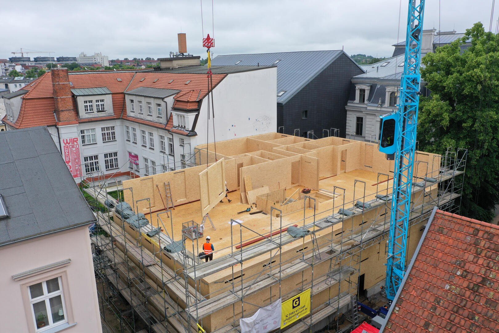 A construction site shows a multi-storey wooden building surrounded by scaffolding. Workers and a blue crane can be seen. The construction site is surrounded by older buildings and green areas under a cloudy sky.