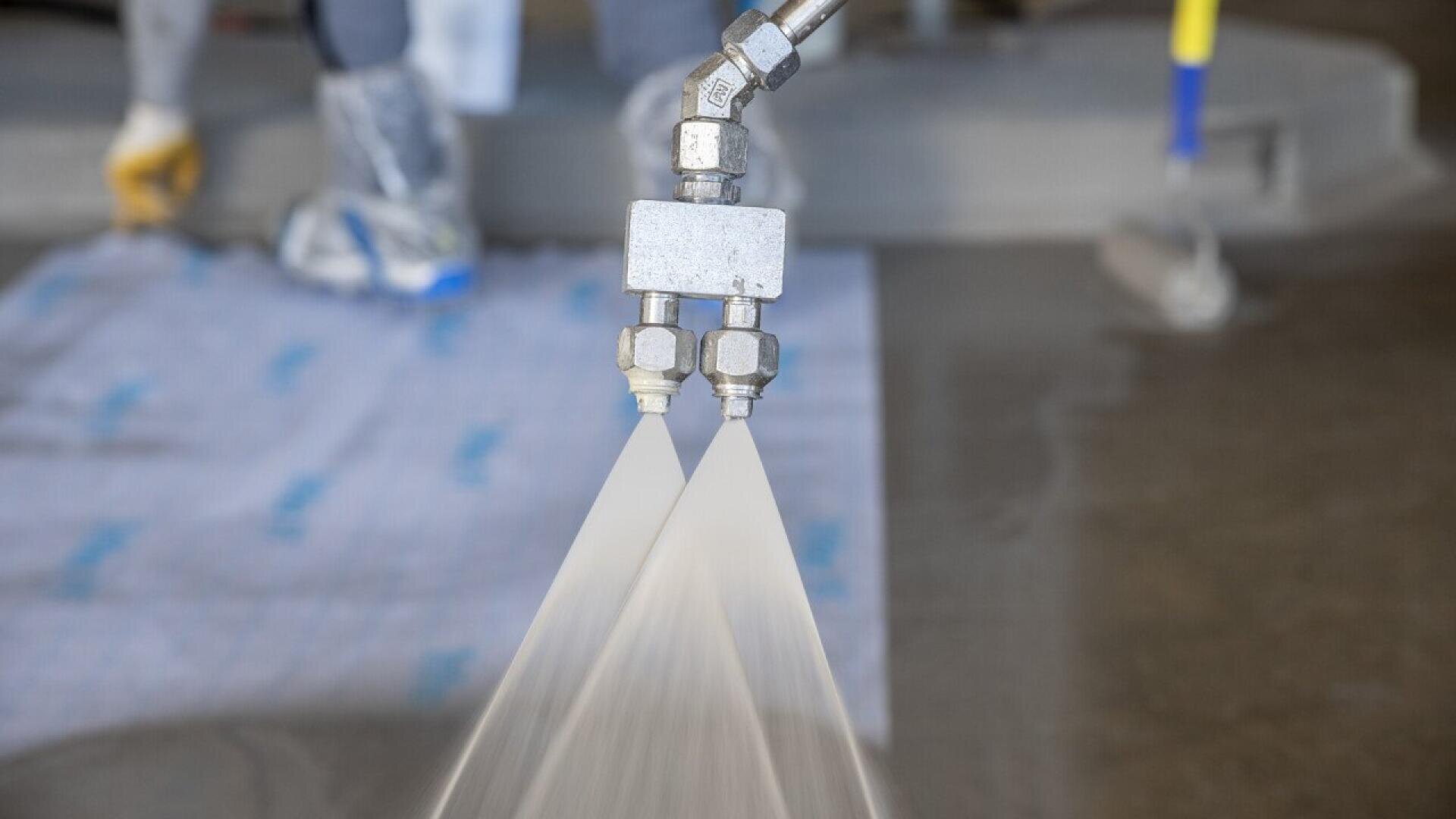 Close-up of a dual-nozzle sprayer releasing two streams of liquid onto a surface, with industrial flooring and workers feet visible in the blurred background.