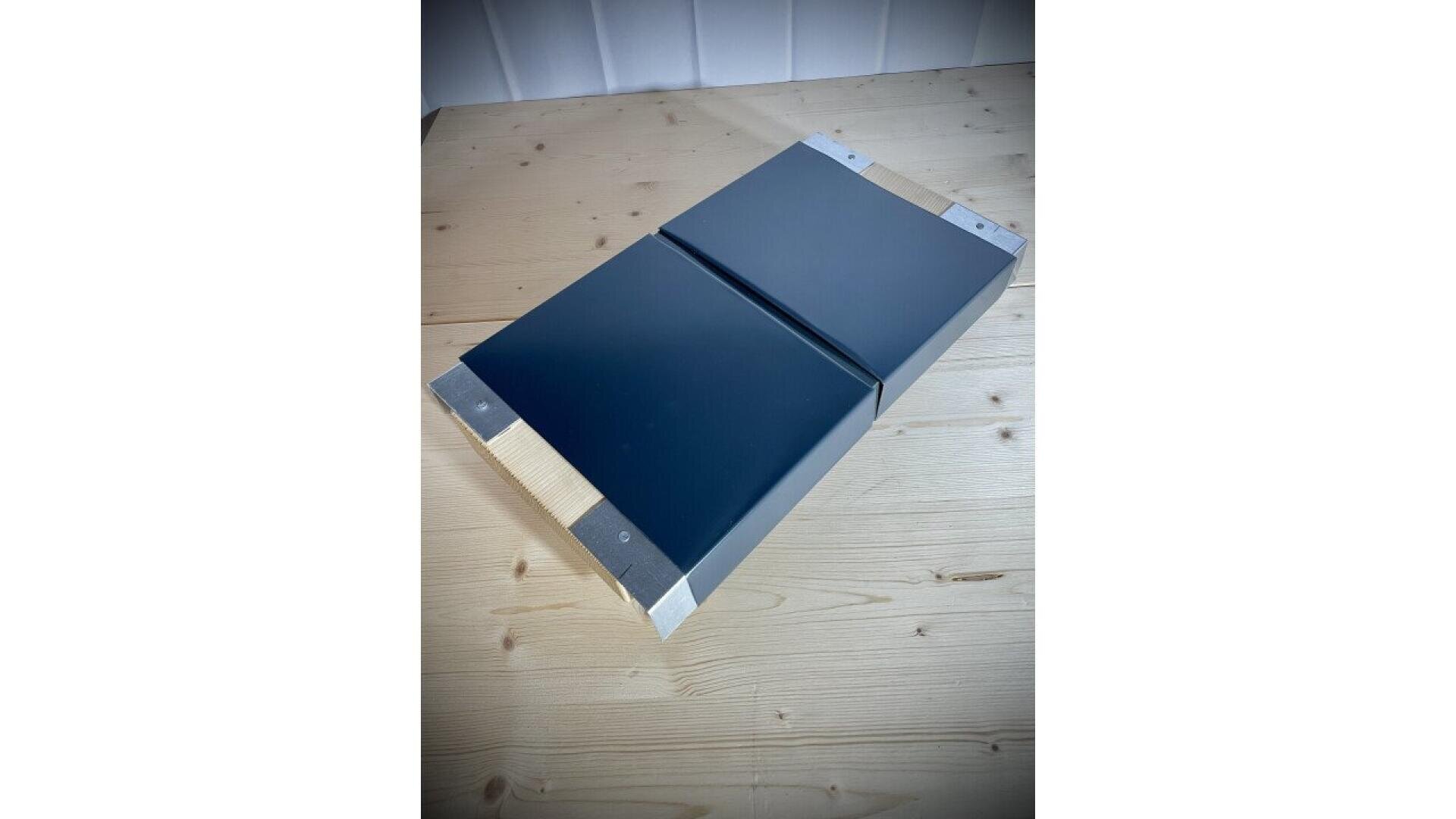 A rectangular black object with metallic edges resting on a light wooden surface; it appears to have a seam down the center, possibly a folding or hinged feature.