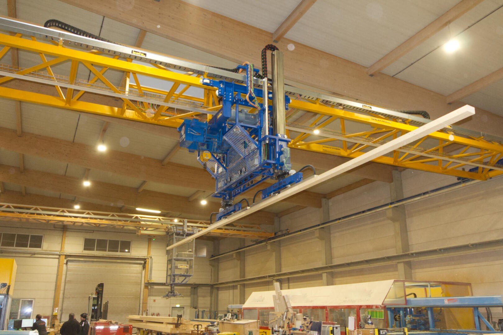 Eine große blau-gelbe Industriemaschine bewegt mit einem Vakuumheber eine lange weiße Platte in einer geräumigen Fabrik mit hohen Decken und sichtbaren Balken.