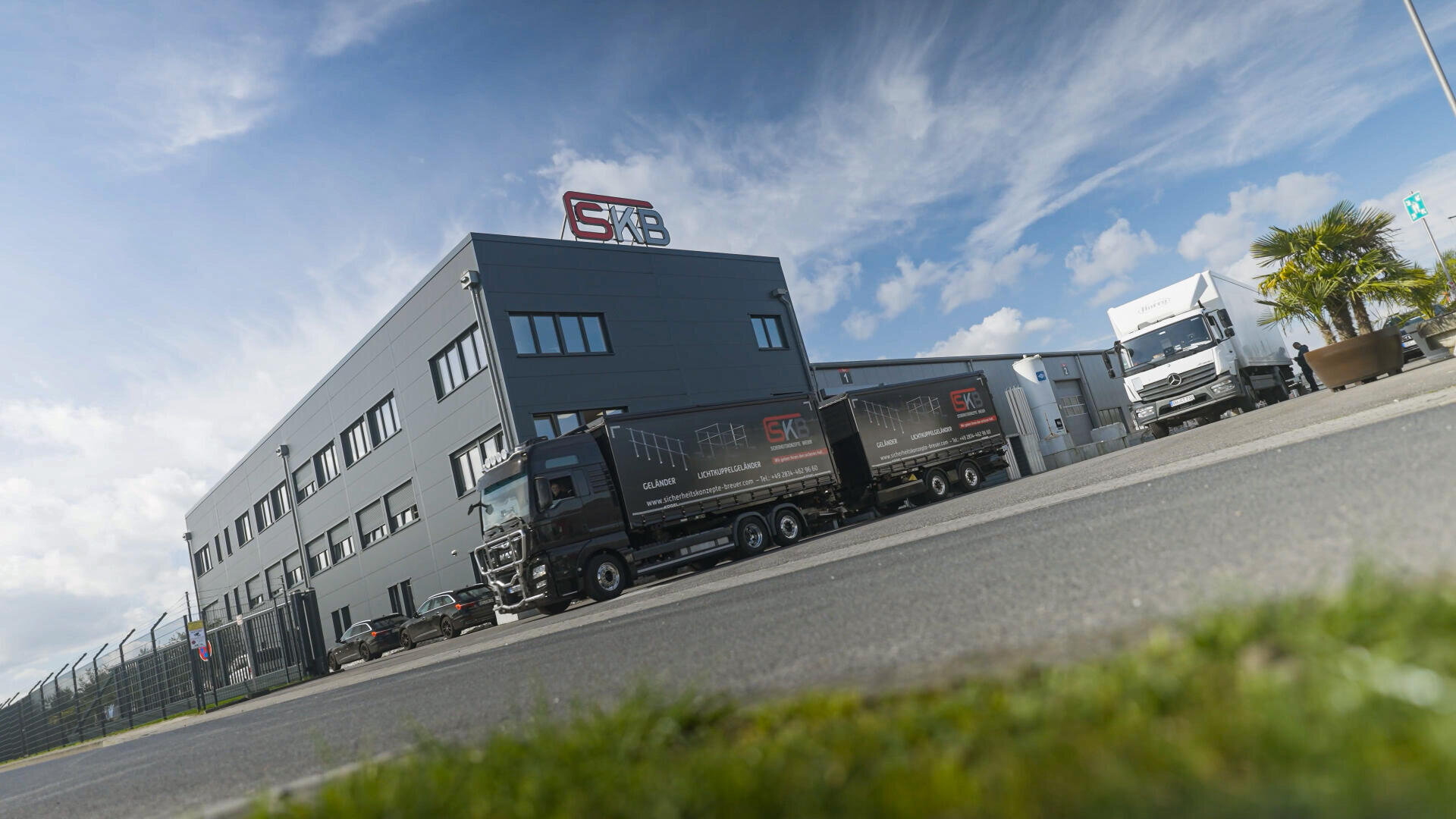 Modernes Industriegebäude mit dem Logo der SFB auf dem Dach, zwei schwarze LKWs davor, ein weißer LKW im Hintergrund und eine Palme rechts. Der Himmel ist teilweise bewölkt.