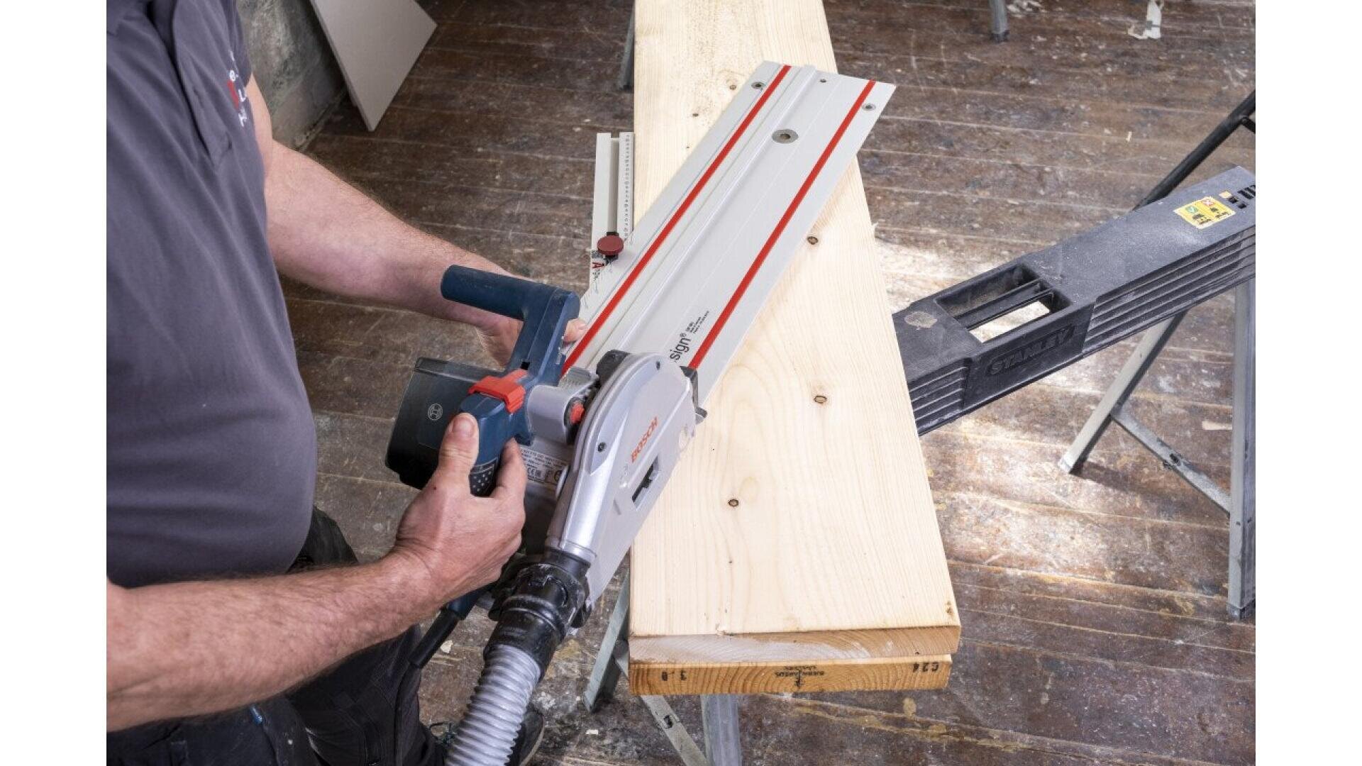 Eine Person schneidet mit einer Tauchsäge ein helles Holzbrett auf einer Werkbank zu. Auf dem Boden ist Sägemehl zu sehen und in der Nähe liegt ein weiteres Werkzeug.