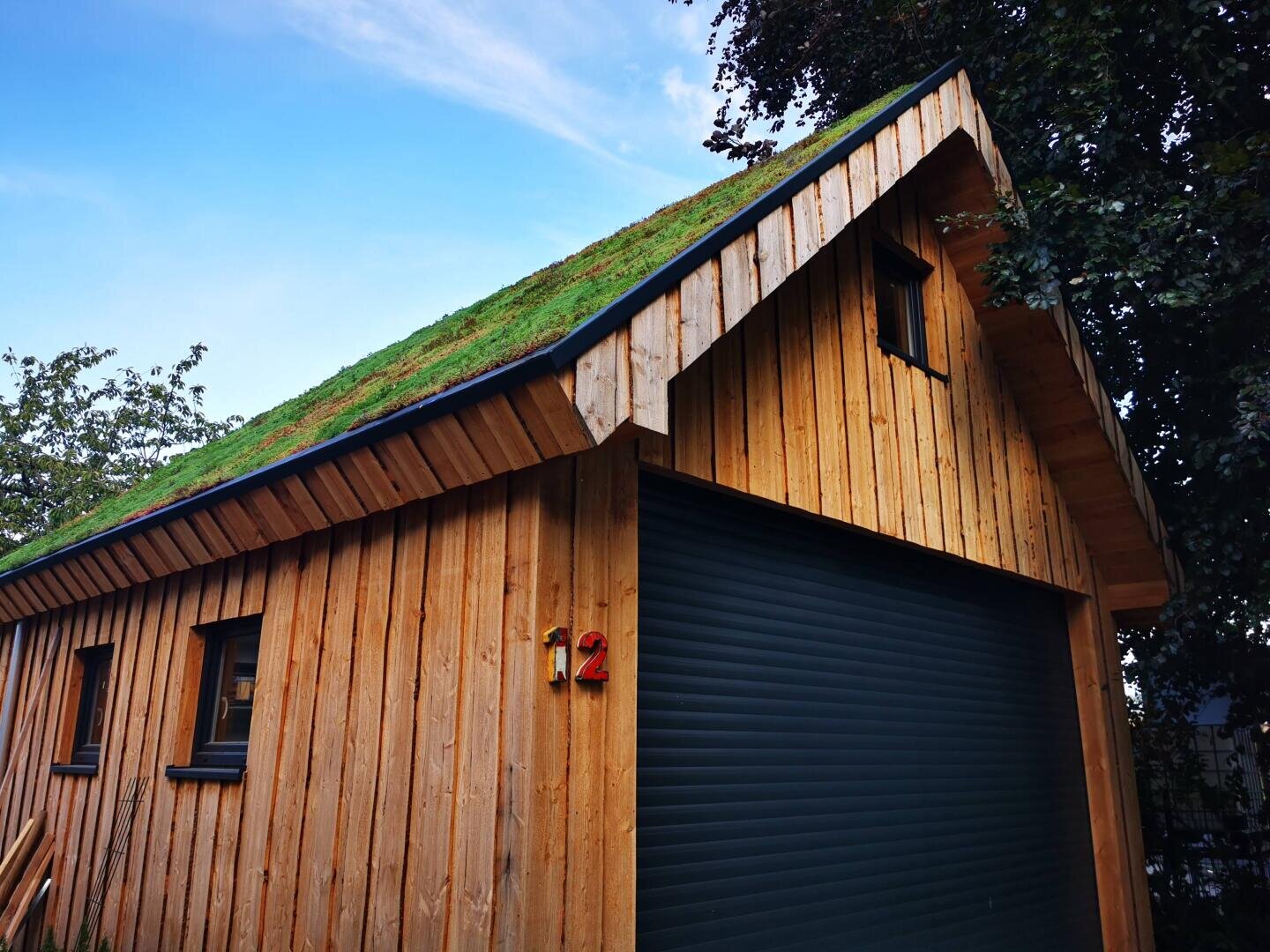 Eine Holzgarage mit grünem, grasbedecktem Dach, einem kleinen Fenster darüber, einem großen schwarzen Rolltor und der Zahl 12 in der Nähe des Eingangs, umgeben von Bäumen unter einem blauen Himmel.