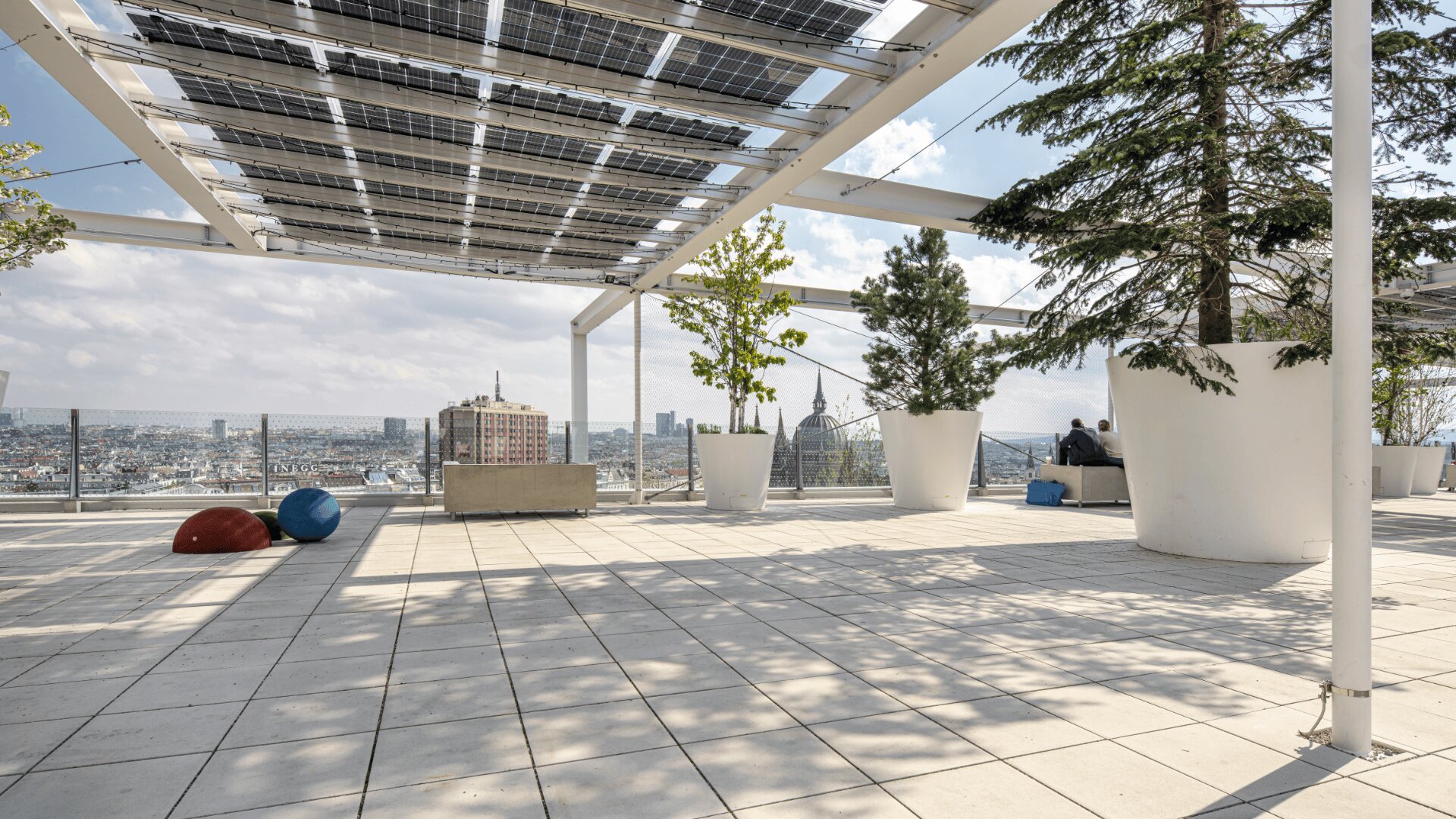 Eine großzügige Dachterrasse mit großen Topfbäumen, Solarpanelen und Blick auf die Stadt. Im Hintergrund sitzen zwei Personen auf einer Bank; bunte runde Sitze stehen auf dem gefliesten Boden unter einem leicht bewölkten Himmel.