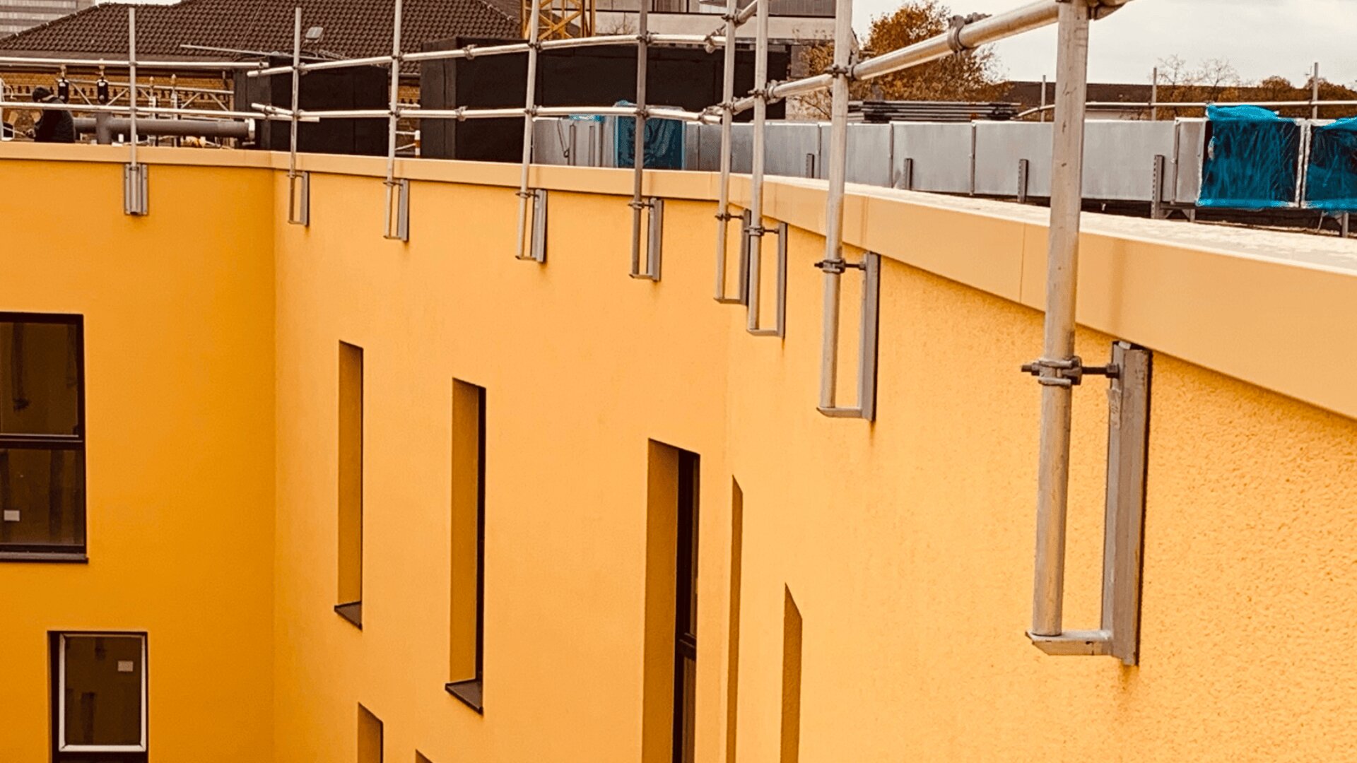 Ein gelbes Gebäude mit vertikalen, rechteckigen Fenstern und Metallgeländern an der Dachkante. Im Hintergrund sind Baumaterialien und ein weiteres Gebäude zu sehen.
