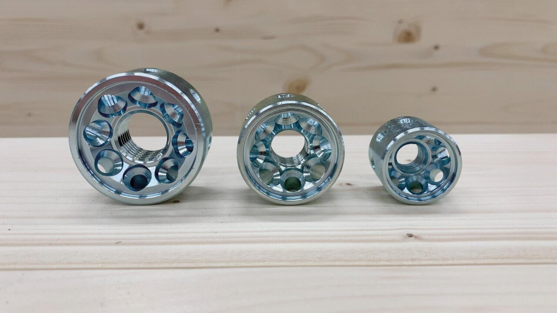 Three shiny metal components of varying sizes are arranged in a row on a light wooden surface with a light wood background, showing circular shapes and multiple holes in each piece.