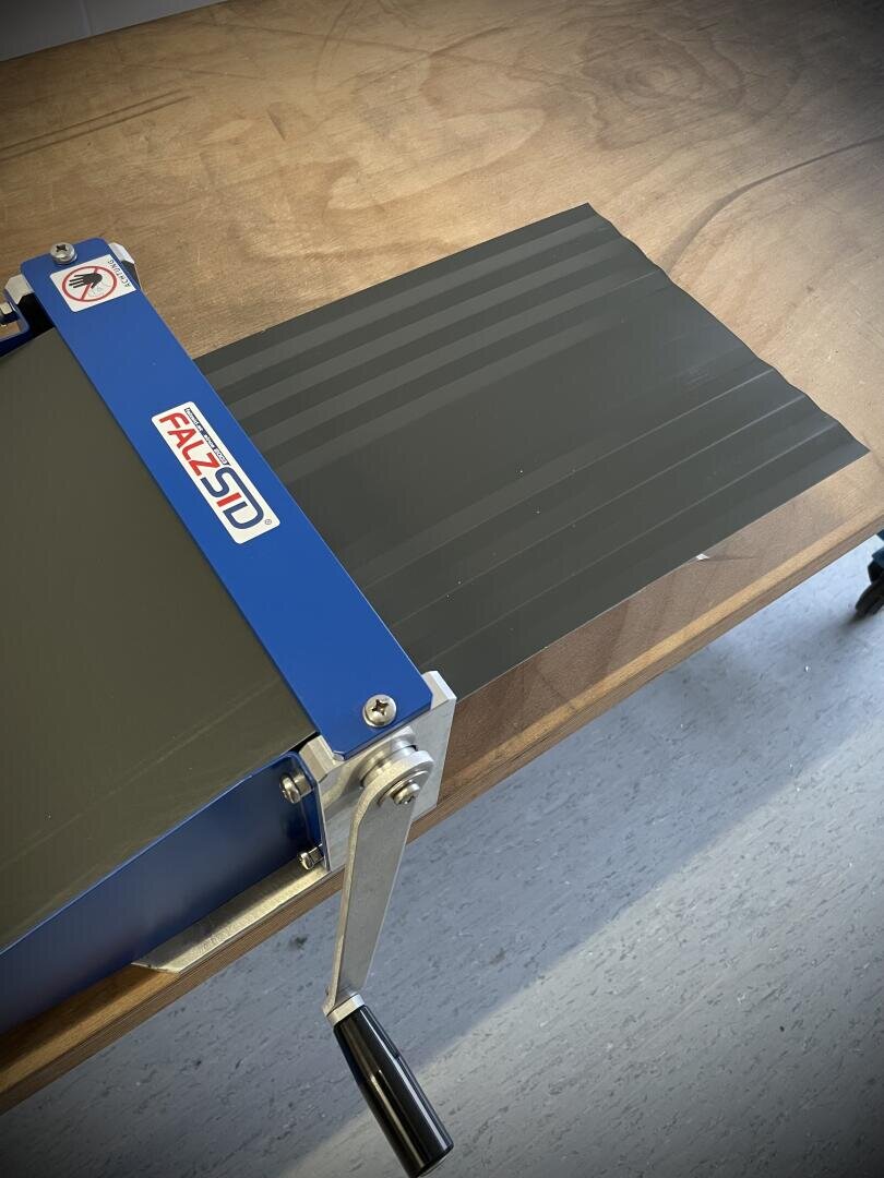 Eine blau-silberne Handkurbelmaschine auf einem Holztisch erzeugt parallele Prägelinien auf einem schwarzen Materialblatt. Die Maschine trägt die Bezeichnung FALZ3D und verfügt über eine Kurbel zur manuellen Bedienung.