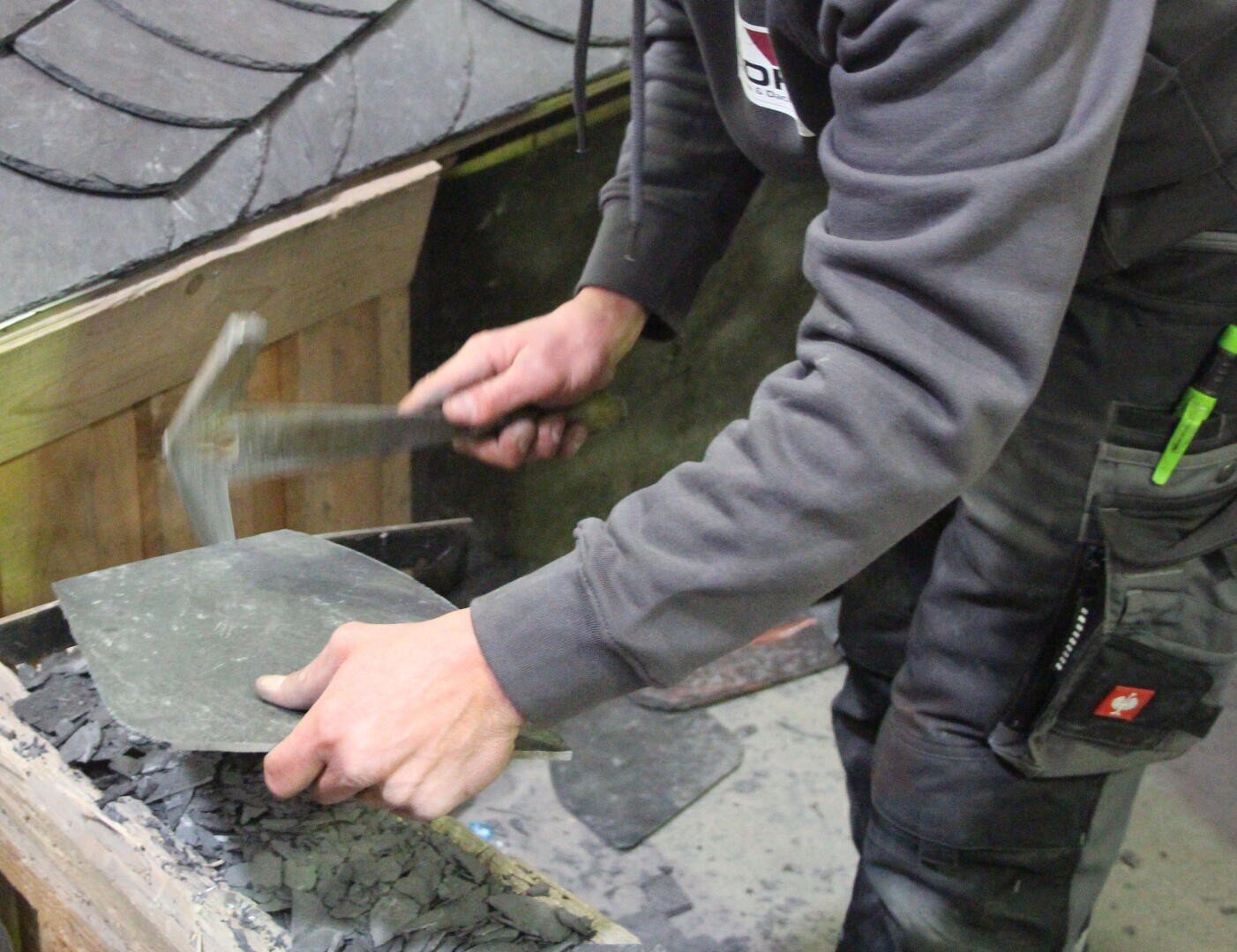 Eine Person in Arbeitskleidung formt mit einem Hammer ein Stück Schiefer, wahrscheinlich für die Dacheindeckung, während sie ihn über eine mit Schieferbruchstücken bedeckte Fläche hält.
