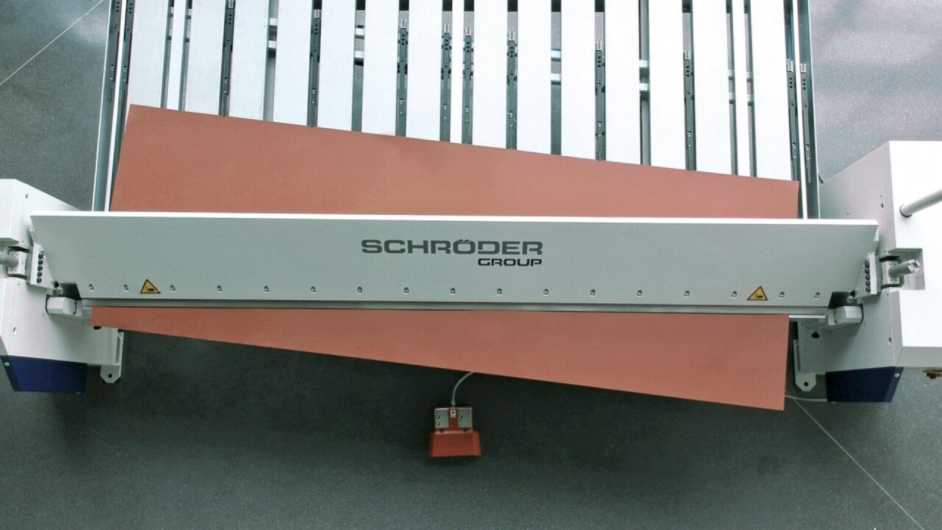 A sheet metal bending machine from Schröder Group is bending a large brown metal sheet, with the bent sheet extending beyond the edge of the machine and a red foot pedal visible below.