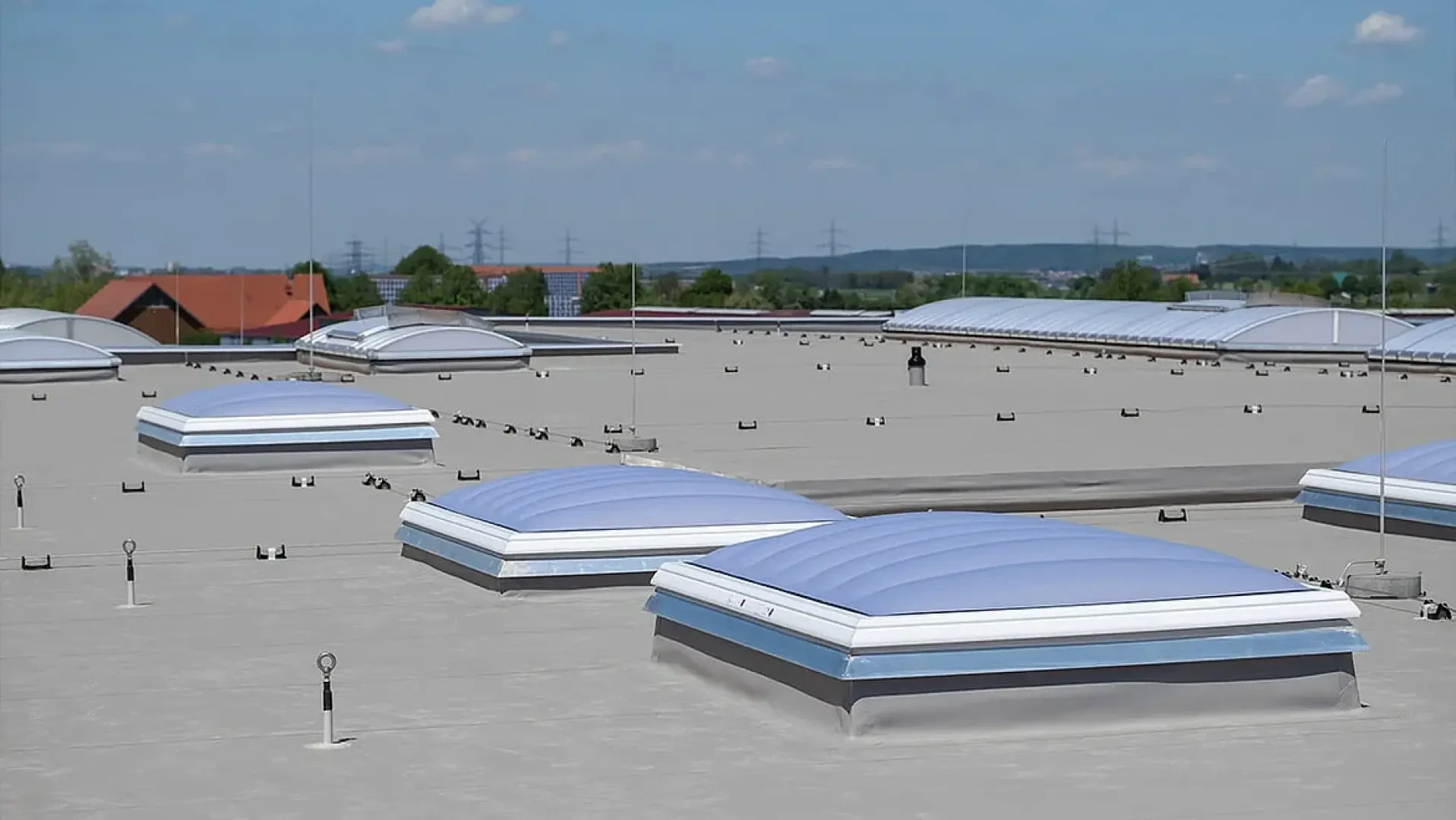 Flaches Dach mit mehreren großen, gleichmäßig verteilten kuppelförmigen Oberlichtern. Der Hintergrund zeigt entfernte Bäume, Häuser und Stromleitungen unter einem blauen Himmel mit vereinzelten Wolken.