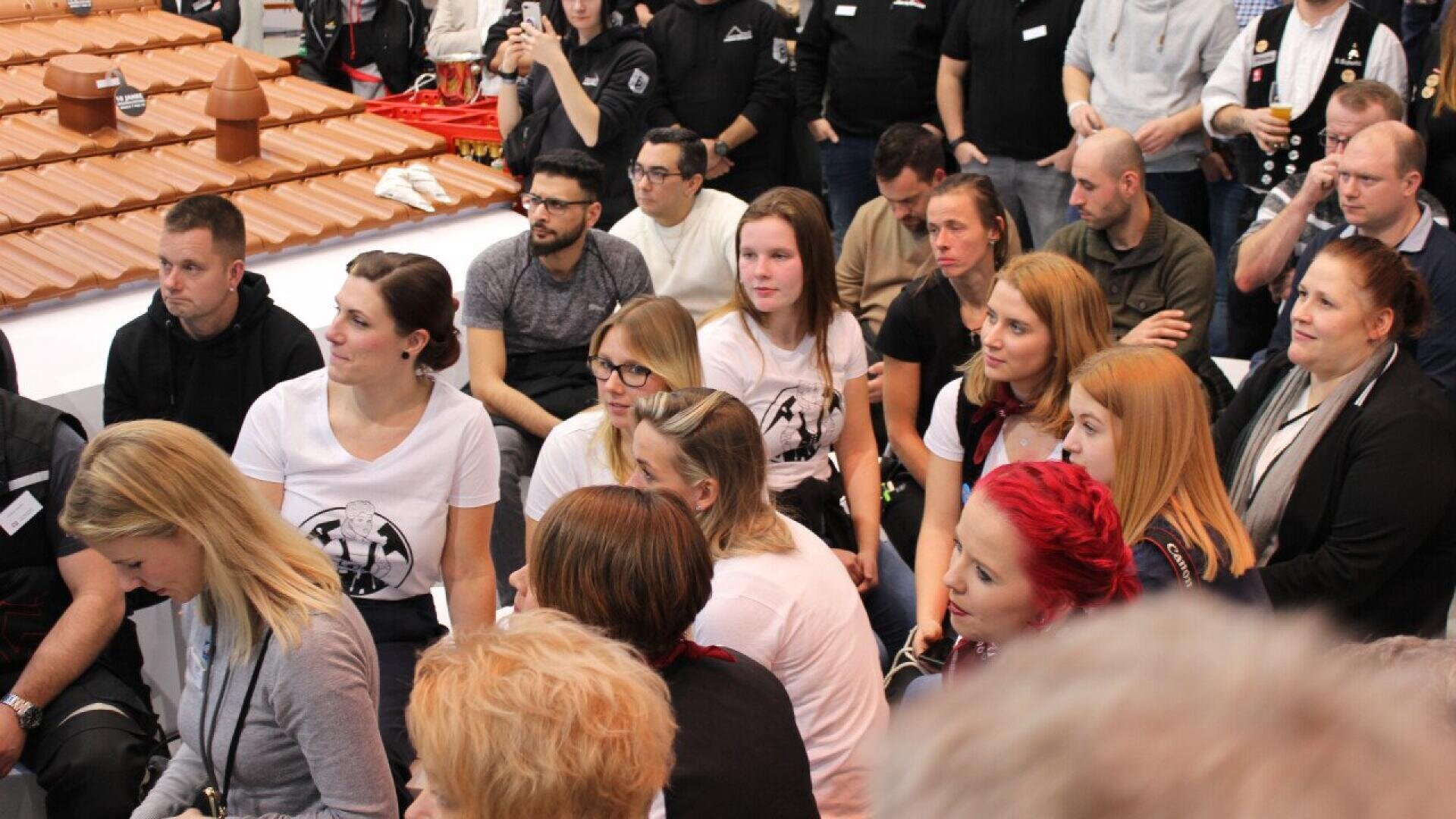 A group of people, mostly women, sit closely together indoors, attentively watching something out of frame. Some wear matching white shirts with a black graphic. In the background, there’s a display with small model rooftops.