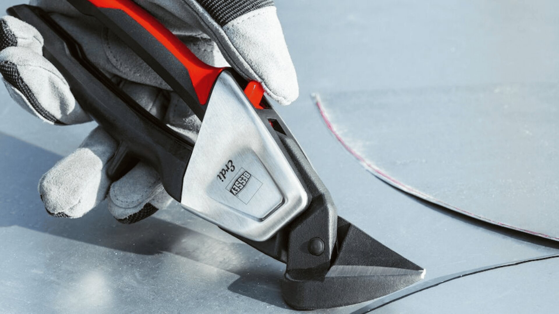 Eine behandschuhte Hand schneidet mit einer Metallschere mit schwarz-rotem Griff entlang einer gekrümmten Linie durch ein Blech.