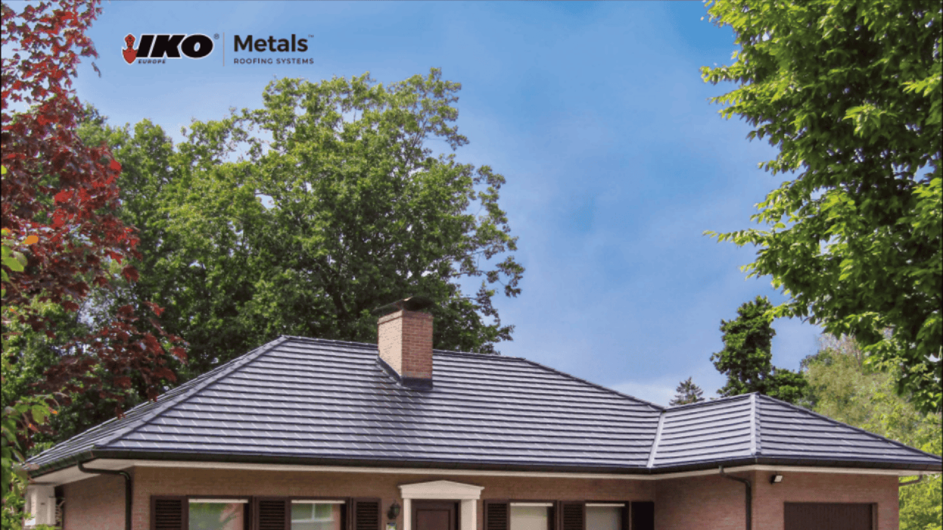 A house with a dark metal tile roof sits beneath a clear blue sky, surrounded by green and red-leaved trees. The IKO Metals Roofing Systems logo appears in the top left corner.