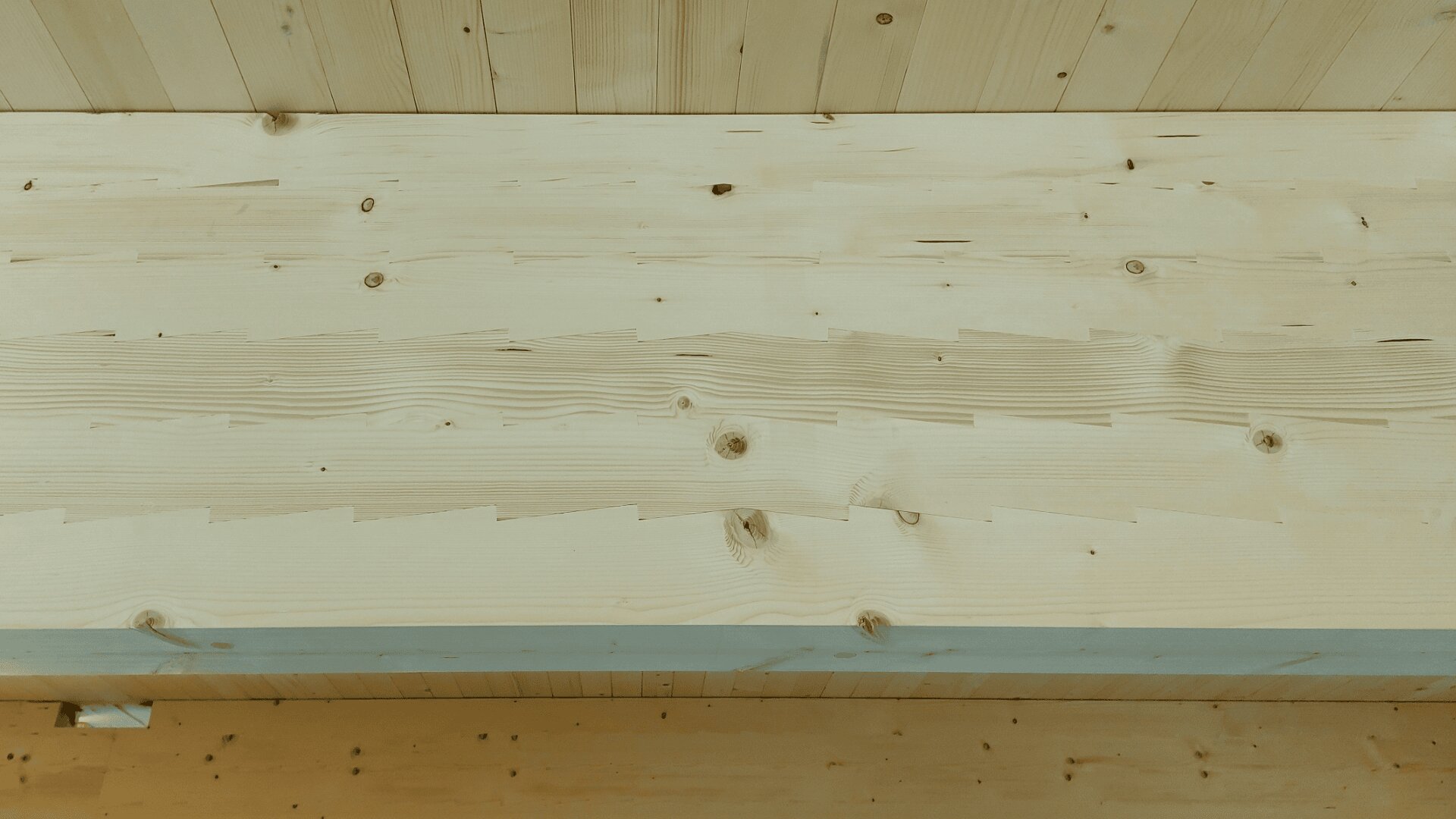 Nahaufnahme einer Decke aus hellen Holzbrettern mit sichtbaren Knoten, Maserungen und Nähten, die ein natürliches und strukturiertes Aussehen verleihen.