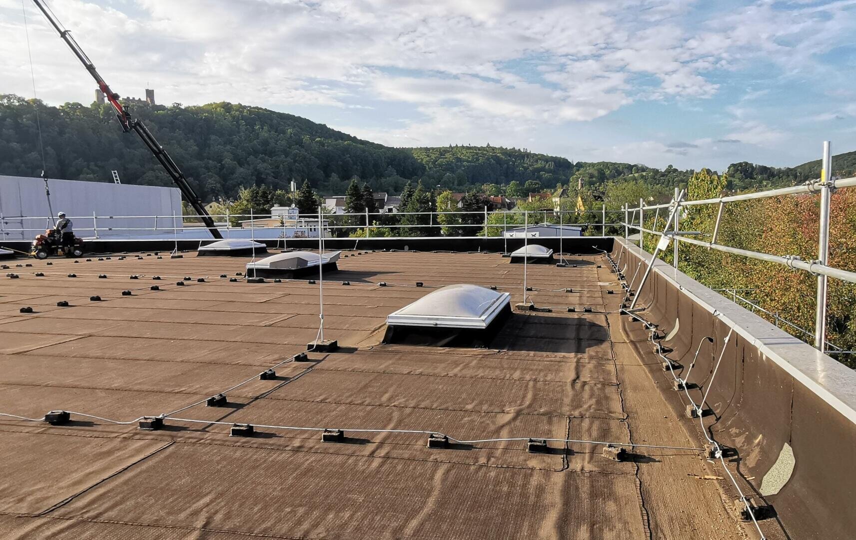 Ein flaches Dach mit Sicherheitsgeländern, mehreren kuppelförmigen Oberlichtern und isolierten Paneelen mit Blick auf Bäume und grüne Hügel unter einem teilweise bewölkten Himmel. Im Hintergrund sind Baumaschinen und ein Kran zu sehen.