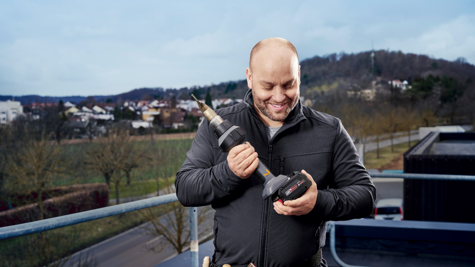 Ein lächelnder Mann in einer schwarzen Jacke steht draußen auf einem Balkon. Er hält eine Akkubohrmaschine in der Hand und setzt den Akku ein. Im Hintergrund sind Bäume, Gebäude und Hügel unter einem bewölkten Himmel zu sehen.