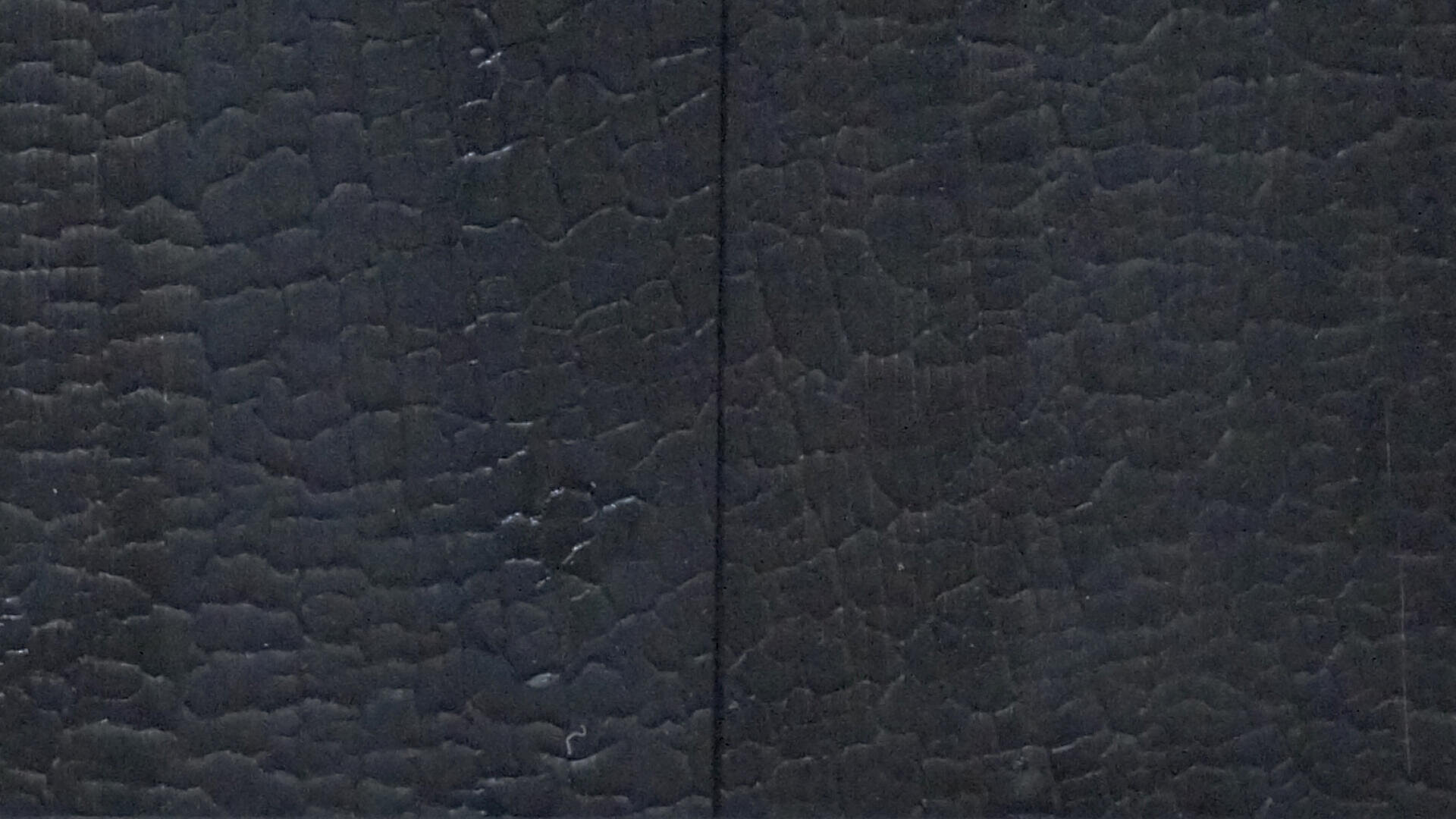 Nahaufnahme von zwei nebeneinander liegenden dunklen Holzbrettern mit einer strukturierten, rissigen und verkohlten Oberfläche, die in der Mitte eine senkrecht verlaufende Naht aufweist. Das Holz scheint für einen dramatischen Effekt verbrannt worden zu sein.