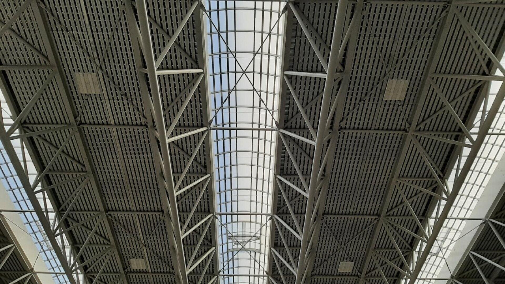 Looking up at a modern industrial ceiling with metal beams, trusses, and panels. Natural light filters through a central skylight, highlighting the geometric structure and grid pattern.