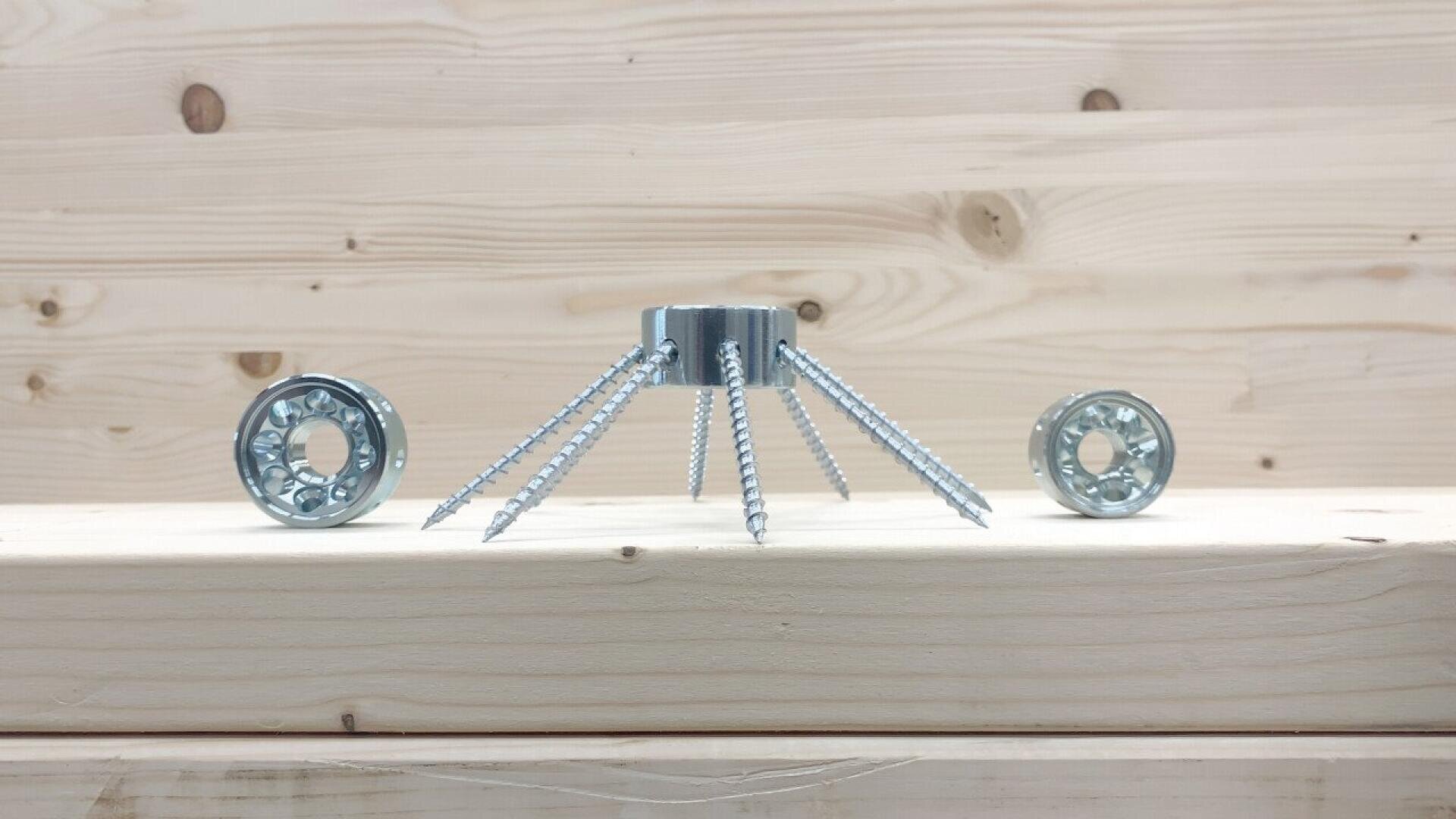 A small metal piece with six screws resembling spider legs is placed upright on a wooden surface, flanked by two round metal parts, all set against a light wood background.