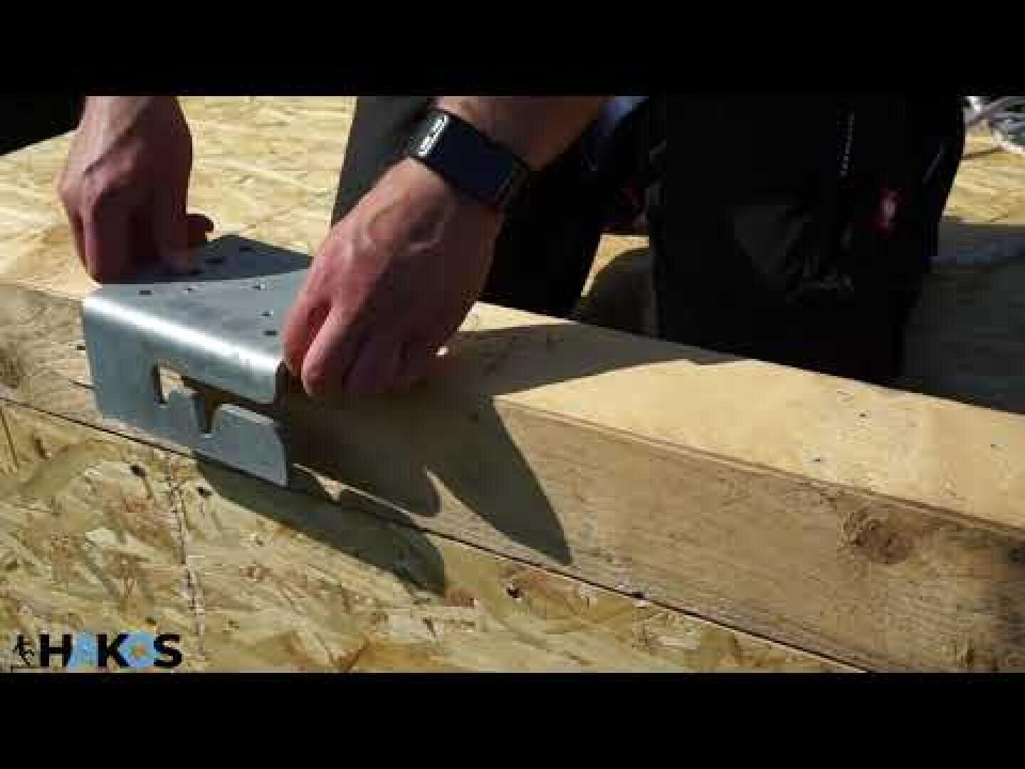 Eine Person, die eine Smartwatch trägt, bringt ein Metallverbindungsstück an einem Holzbalken an und bereitet sich auf den Bau vor. Das Holz hat sichtbare Schrauben, und die Szene wird von natürlichem Tageslicht beleuchtet.