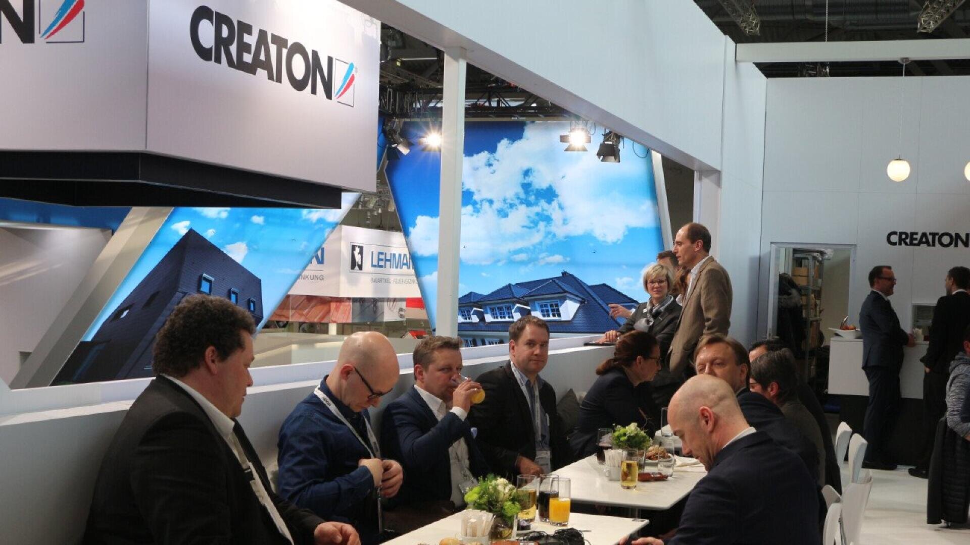 A group of people in business attire sit at a long white table, eating and drinking, at a modern indoor event or trade show. The CREATON brand logo is visible on a large sign above them.