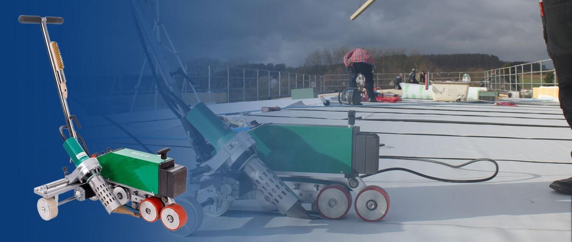 Im Vordergrund ist ein grün-silbernes Schweißautomat mit roten Rädern zu sehen, während im Hintergrund Arbeiter unter bewölktem Himmel Dachmaterialien auf einem Flachdach montieren.