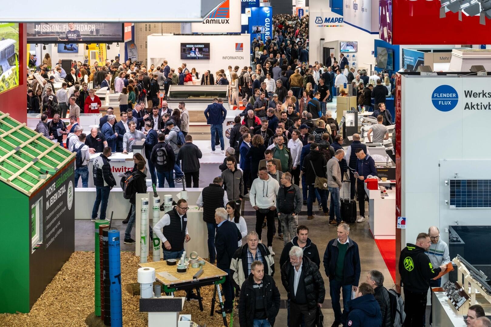 A large crowd of people walk through a busy exhibition hall filled with stands, displays and information signs at a trade fair or congress. Many are engrossed in conversation or looking at the exhibits.