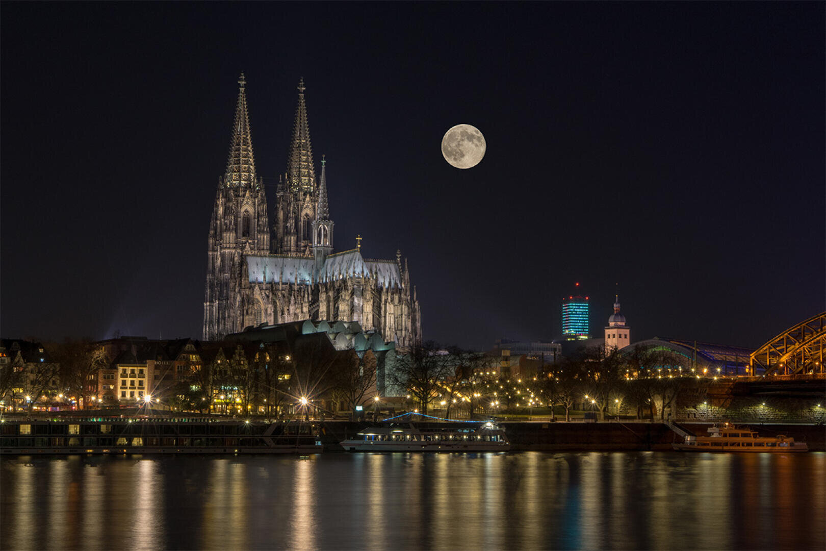 The illuminated Cologne Cathedral stands on the banks of the Rhine at night, with a full moon shining above it. The lights of the city are reflected on the water and a ship is anchored in the foreground.