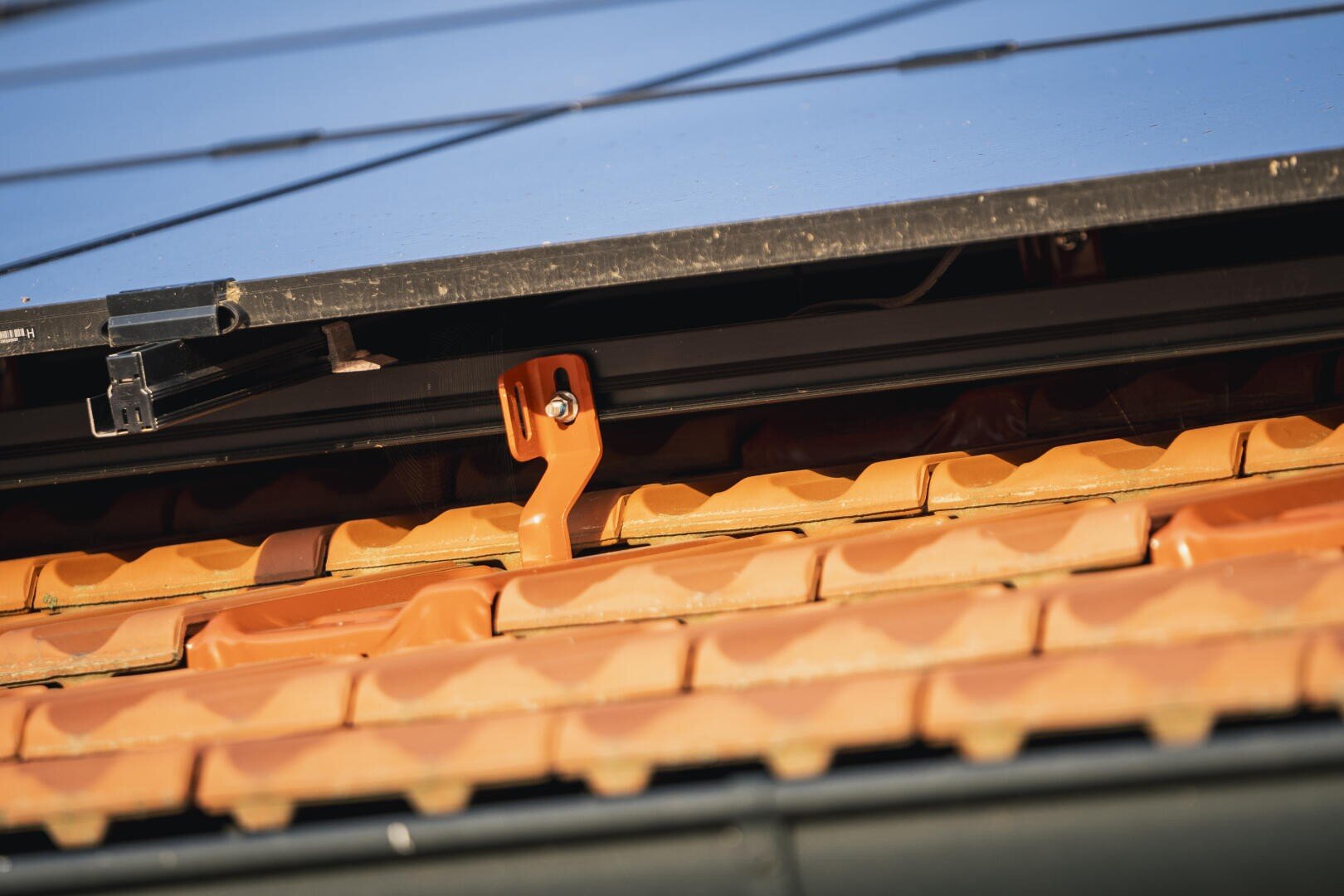 Close-up of a roof with orange-colored tiles, showing the bracket and the underside of a solar module installed above the tiles.
