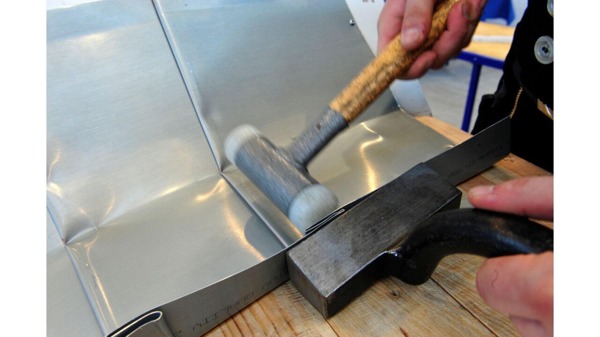 A person uses a wooden-handled mallet and a metal tool to shape or flatten a sheet of metal on a wooden work surface.