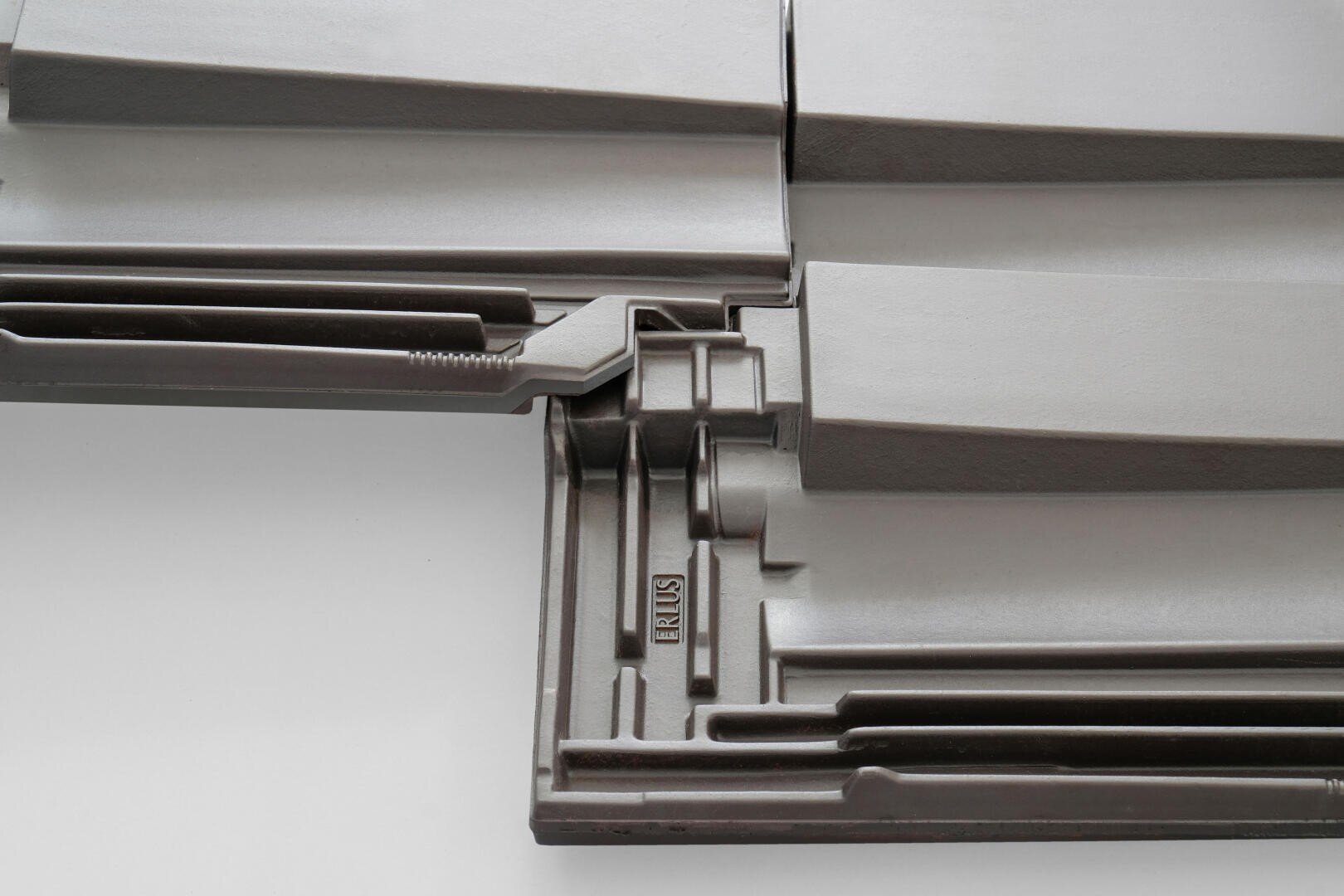 Nahaufnahme ineinandergreifender grauer Dachziegel mit einem strukturierten, geometrischen Muster. Auf einem der Ziegel ist das Wort BRUSS eingeprägt, was auf die Marke oder das Modell hinweist. Die Oberfläche wirkt sauber und glatt.