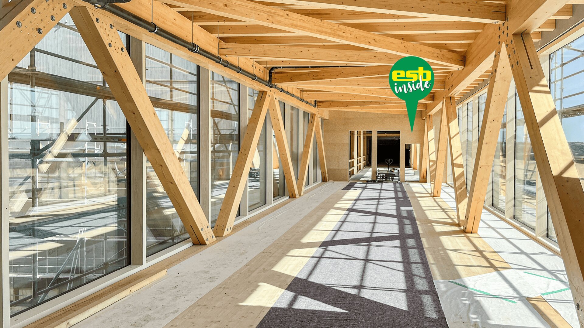 Der lichtdurchflutete Flur mit großen Glasfenstern und freiliegenden Holzbalken schafft eine moderne, offene Atmosphäre. Die Schatten der Balken werfen Schatten auf den Boden. Oben rechts befindet sich ein grünes esp inside-Logo.