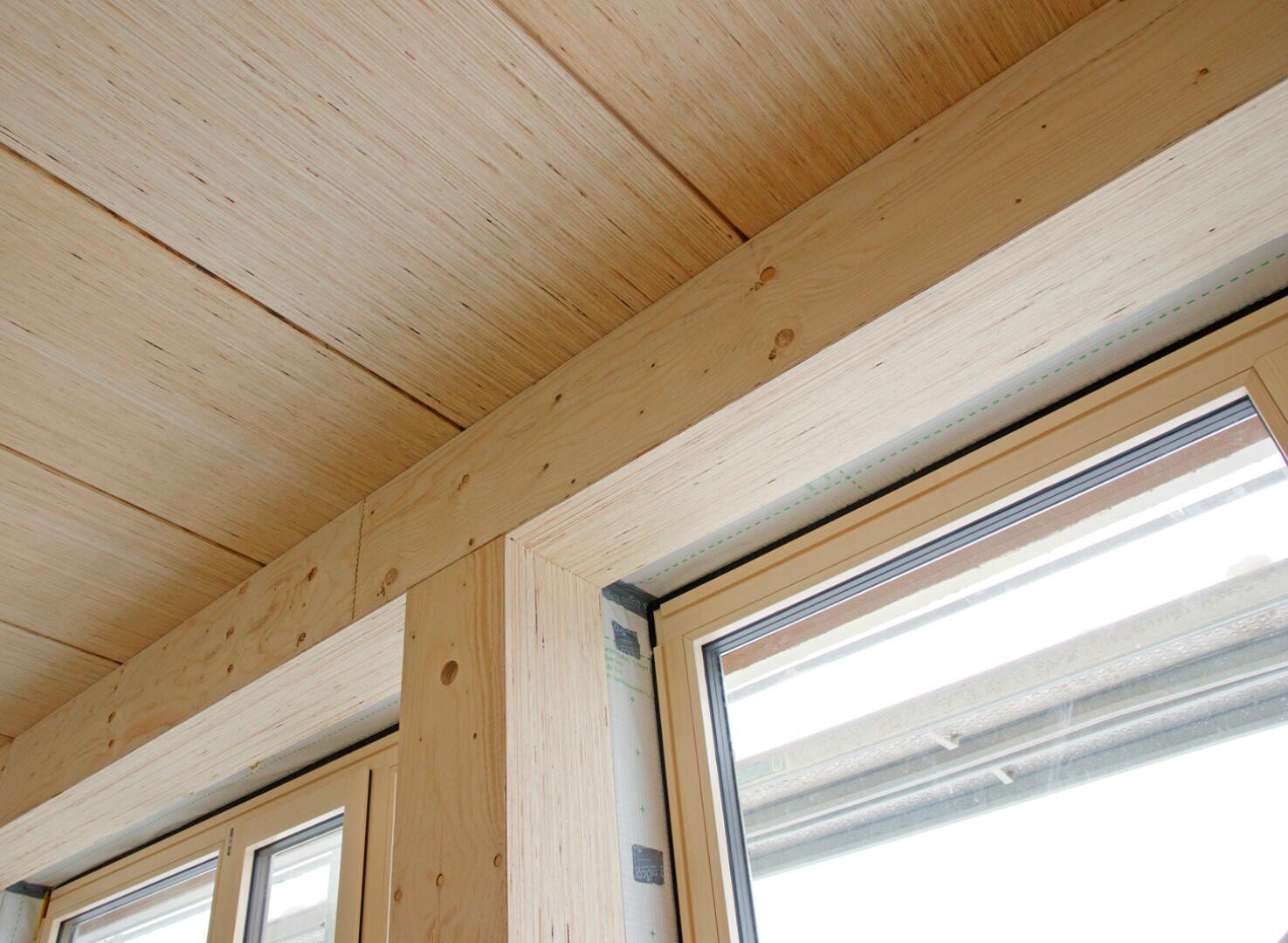 Nahaufnahme einer modernen Holzdecke und eines Fensterrahmens mit hellen Holzpaneelen und Balken sowie großen Fenstern, die natürliches Licht hereinlassen.