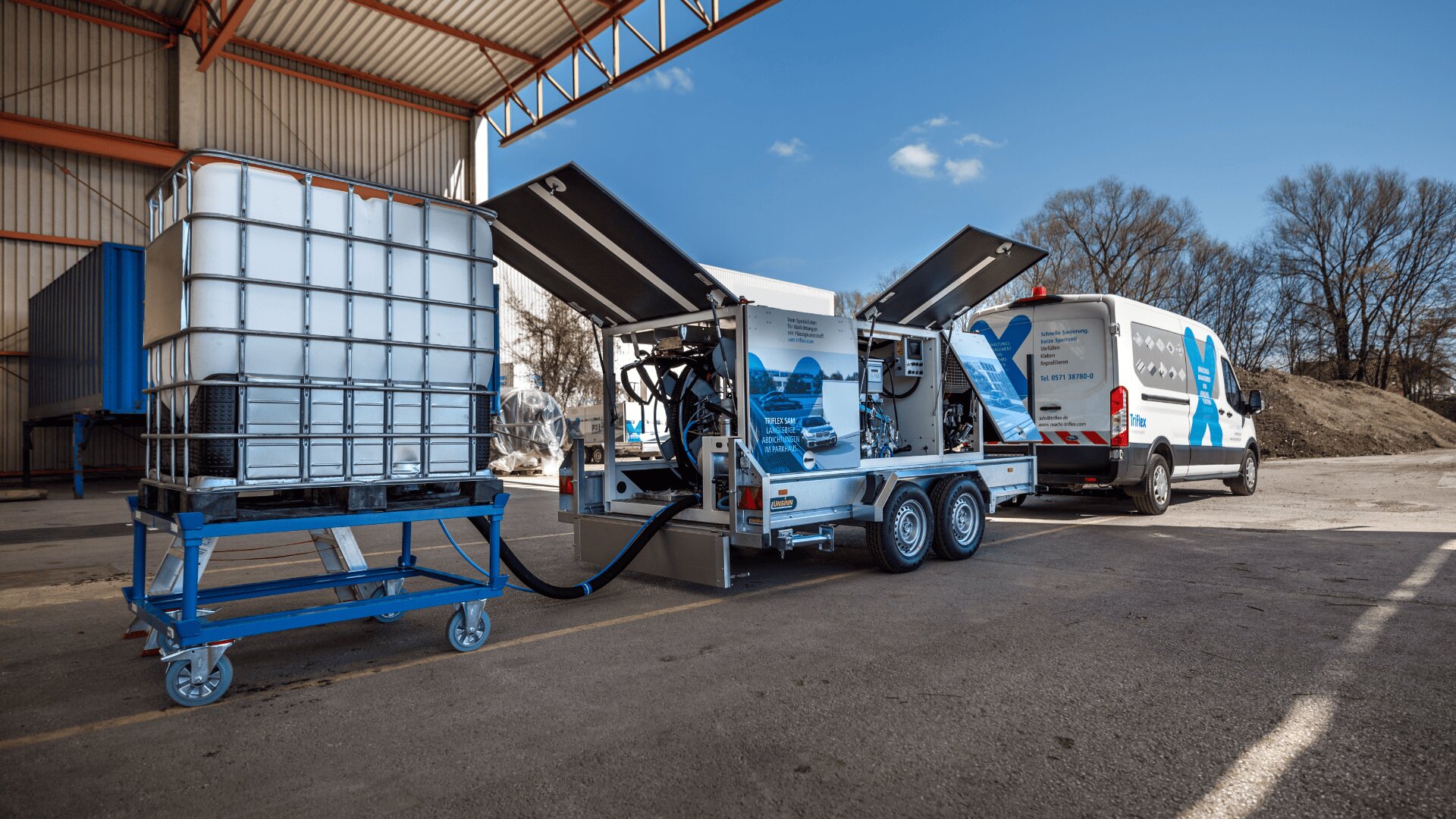 Ein weißer Lieferwagen mit blauer Grafik parkt neben einer mobilen Industriemaschine mit offenen Paneelen und angeschlossenen Schläuchen. Ein großer Wassertank auf einem Metallständer steht in der Nähe in einem Lagerbereich im Freien.