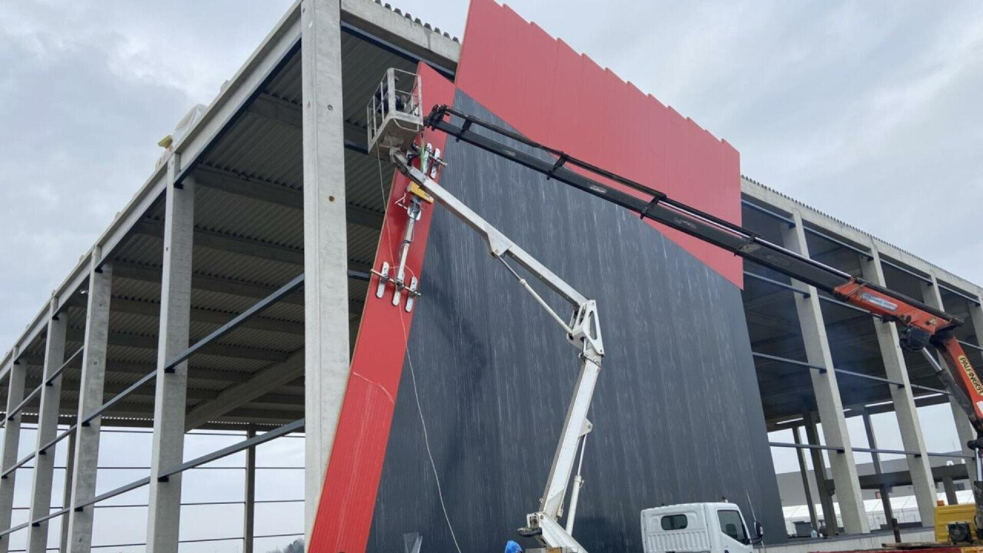Zwei Bauarbeiter bemalen auf hydraulischen Hebebühnen eine große vertikale schwarz-rote Tafel, die an der Seite eines im Bau befindlichen Gebäudes mit Stahlrahmen angebracht ist, während über ihnen ein bewölkter Himmel liegt.
