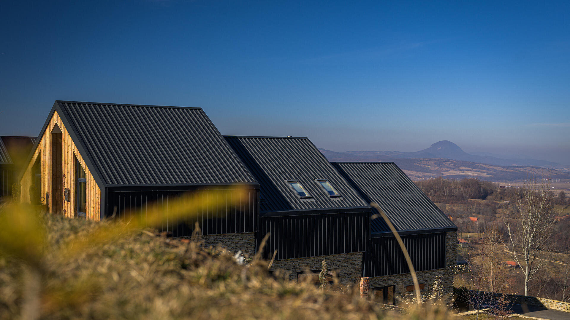 Modern black houses with sloping metal roofs stand on a grassy hill under a bright blue sky, with mountains and valleys in the distance.