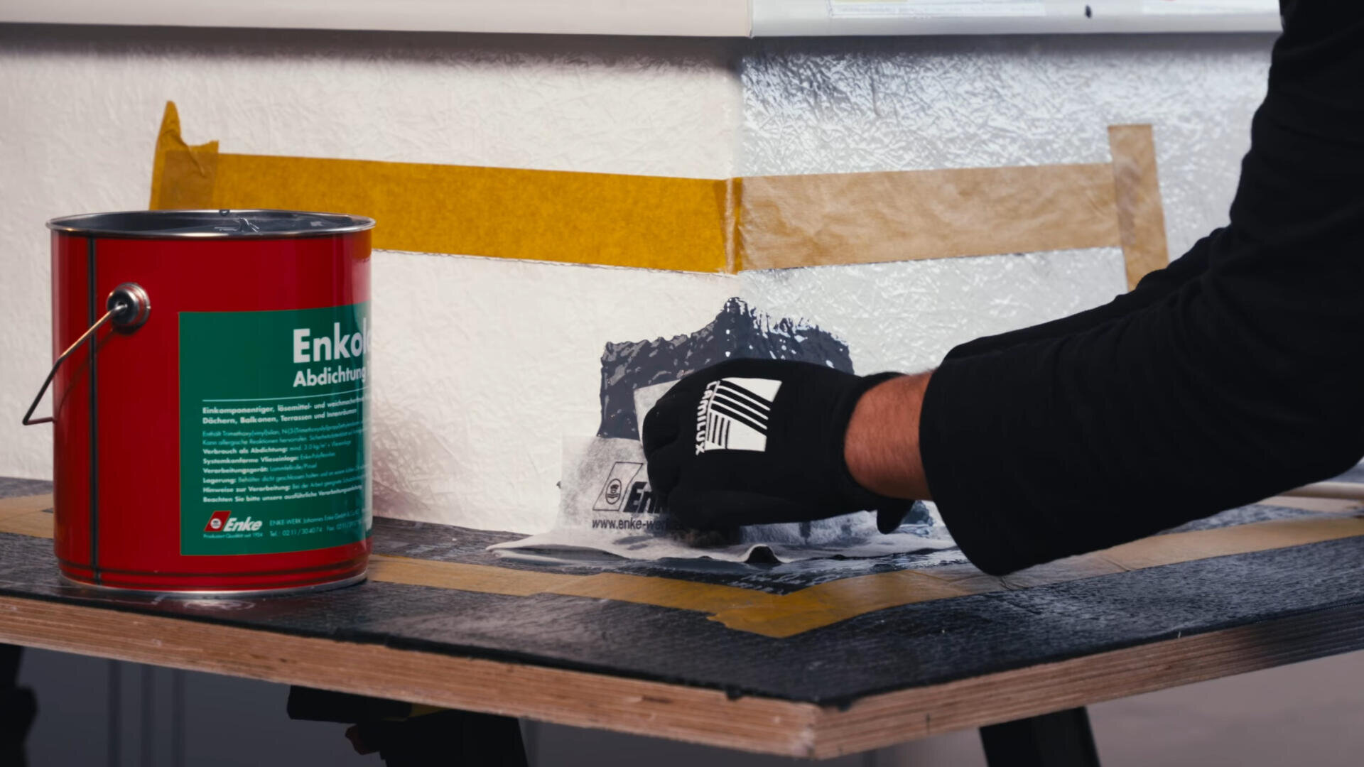 Eine Person mit schwarzen Handschuhen trägt eine dunkle Abdichtungsmasse mit Klebeband auf den Sockel einer Wand auf, daneben steht eine rote Dose Enkopur-Dichtstoff auf einer schwarzen Arbeitsfläche.