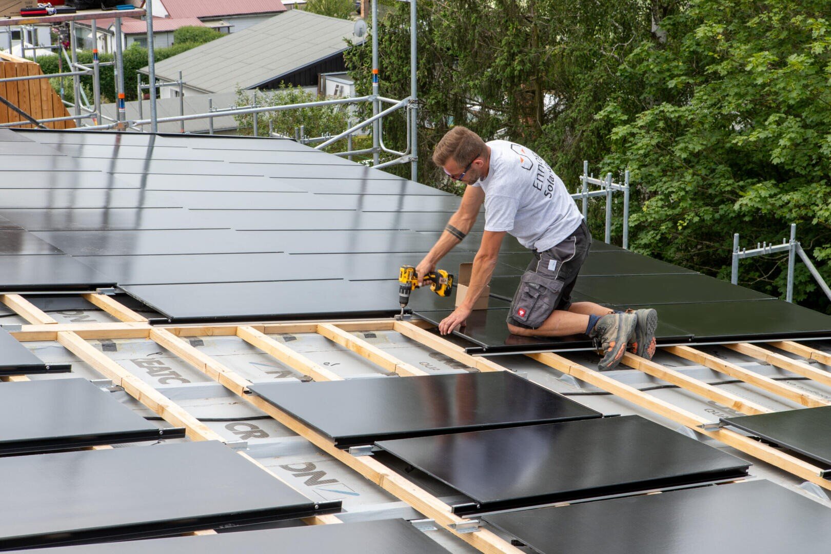Eine Person kniet auf einem schrägen Dach und installiert mit einer Bohrmaschine Solarzellen. Das Holzgerüst stützt die Paneele, und im Hintergrund sind Bäume und Gebäude zu sehen.