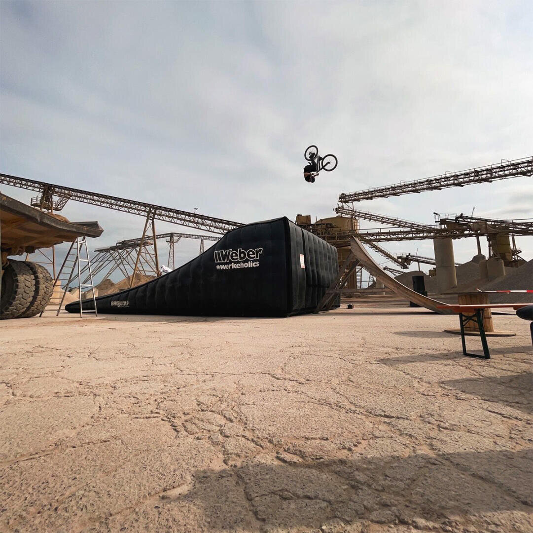 Eine Person auf einem Fahrrad vollführt einen Trick in der Luft über einer aufblasbaren Landebahn in einer Industrieumgebung mit Förderbändern und Maschinen im Hintergrund.
