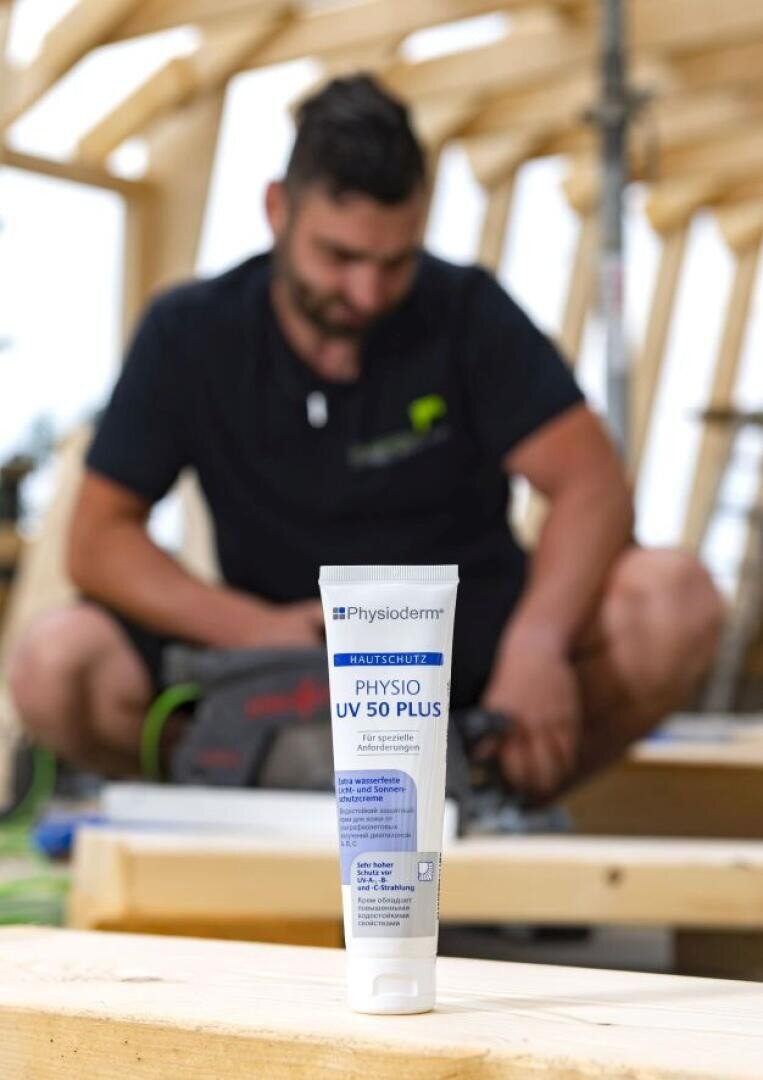 Im Vordergrund ist eine Tube Physioderm UV 50 Plus Sonnenschutzmittel zu sehen, während im unscharfen Hintergrund ein Mann ein Elektrowerkzeug benutzt und Holzkonstruktionen baut.
