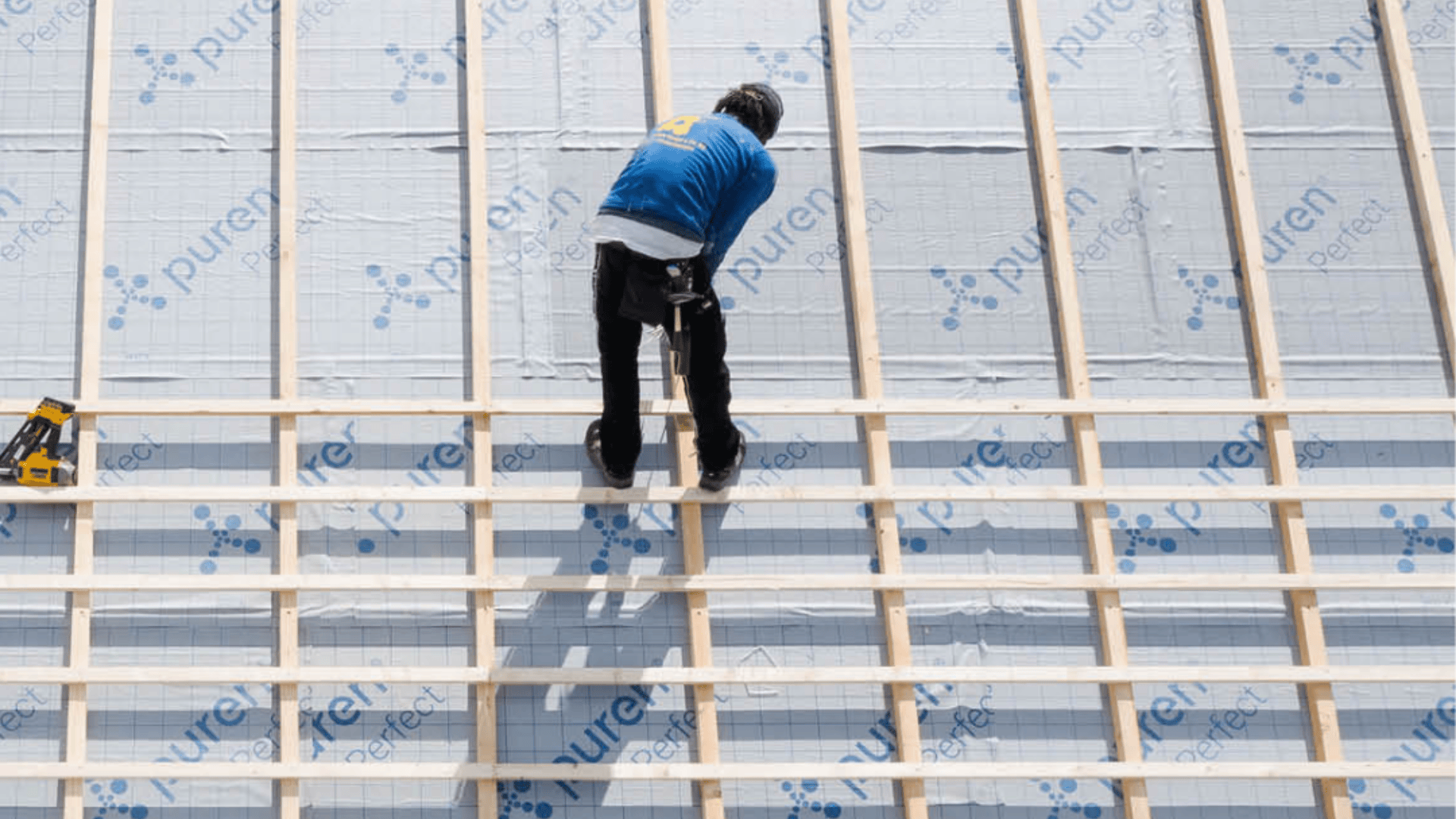 Ein Arbeiter in einer blauen Jacke montiert Holzlatten auf einem mit Dämmplatten gedeckten Dach und bereitet möglicherweise die Montage der Dacheindeckung vor. Die Platten sind mit „puren perfect“ beschriftet.