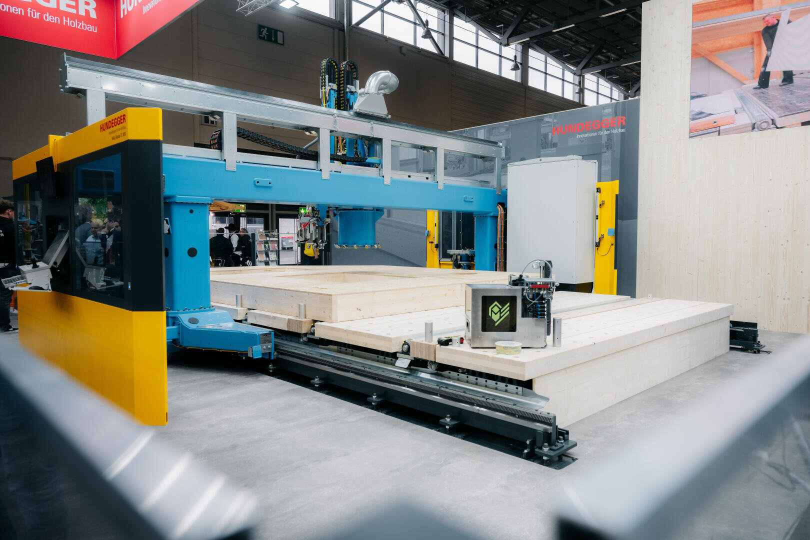 Eine große industrielle CNC-Maschine formt und schneidet Holzplatten auf einer Messe oder in einer Fabrik. Die Maschine ist blau und gelb, auf ihrer Plattform sind fertige Holzplatten ausgelegt. Im Hintergrund sind Menschen und Stände zu sehen.