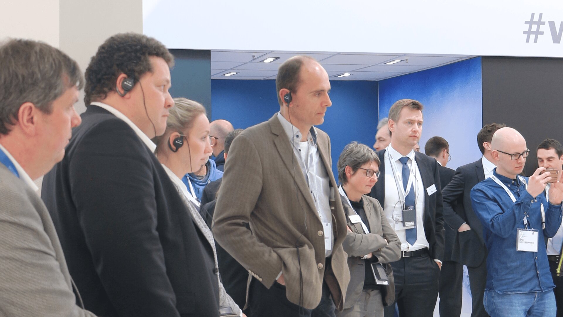 Eine Gruppe von Personen in Geschäftskleidung, einige mit Kopfhörern, steht aufmerksam bei einer scheinbar professionellen Veranstaltung oder Konferenz. Mehrere Personen konzentrieren sich auf etwas außerhalb der Kamera.