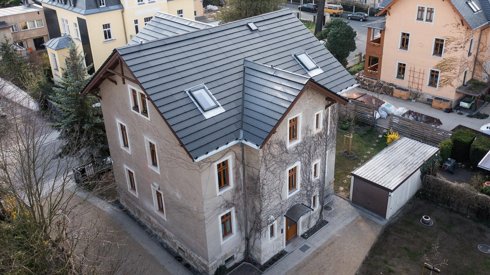 Aerial view of a beige, three-story house with a gray tiled roof, multiple windows, and a small attached garage, surrounded by other residential buildings and trees.